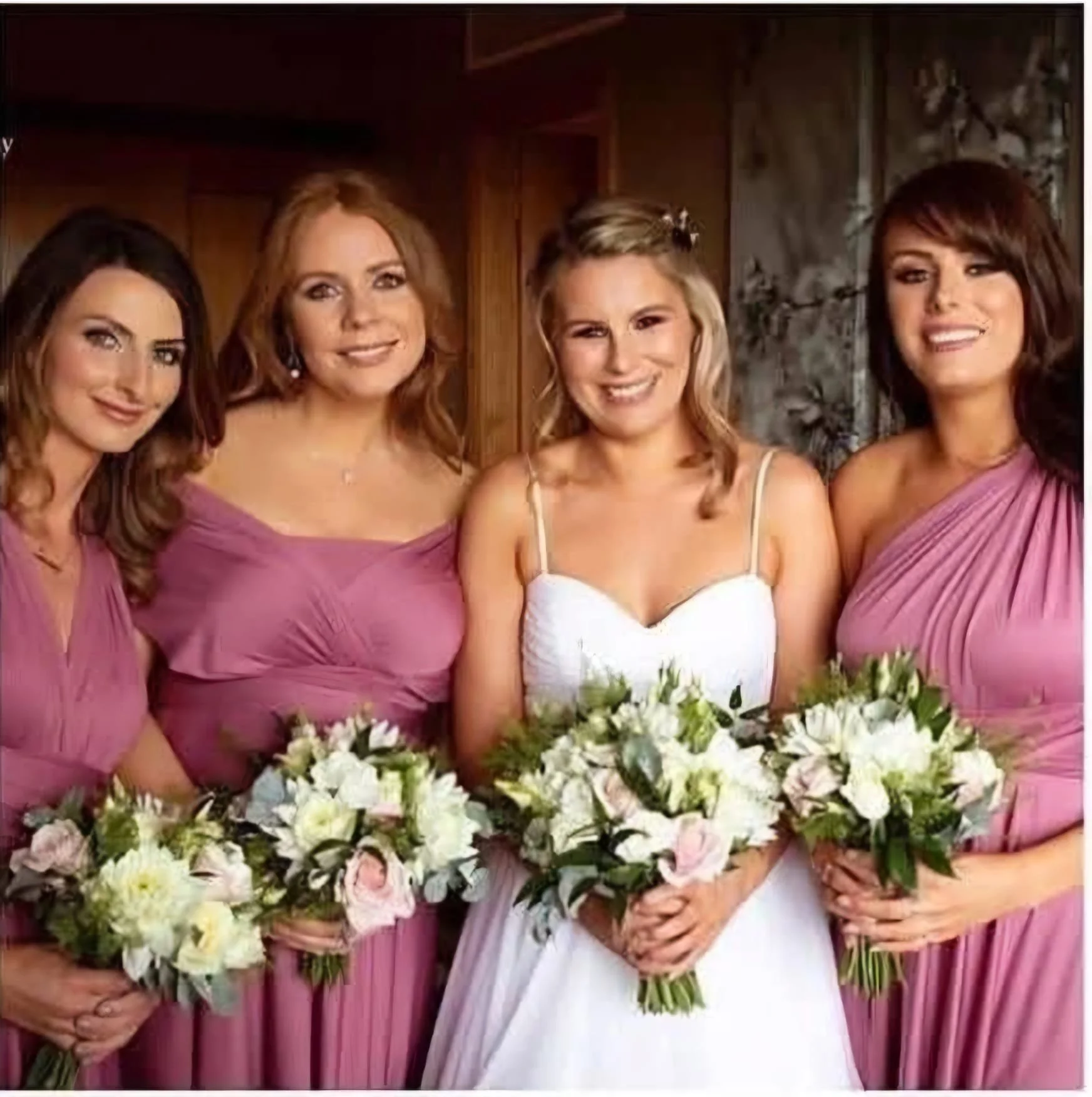 Four women dressed in pink and white formal dresses, holding bouquets of white and pink flowers, standing indoors against a wood and marble background.