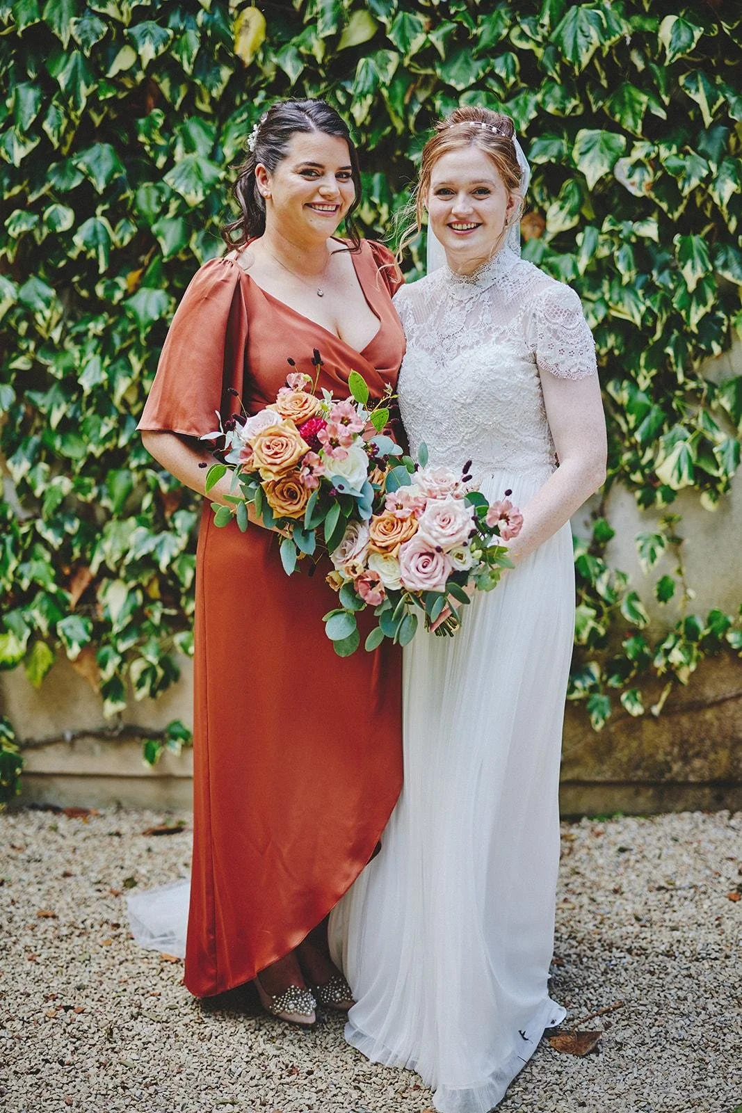 Two women standing side by side outdoors, dressed for a wedding, holding bouquets of pink, peach, and white roses, with ivy-covered wall in the background.