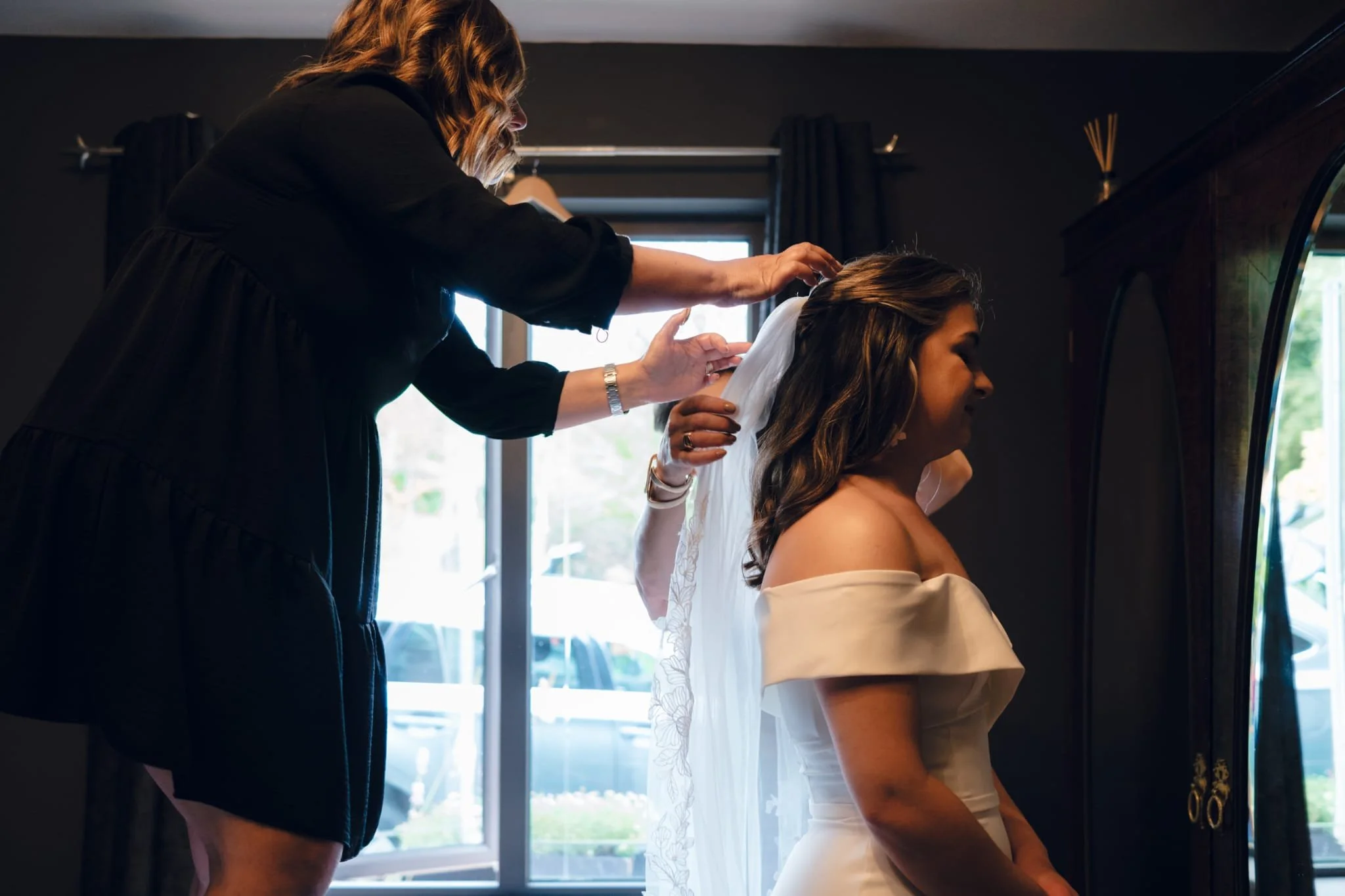 A bride in an off-the-shoulder wedding dress is having her veil adjusted by a woman, possibly her stylist, in a room with dark walls and a window.