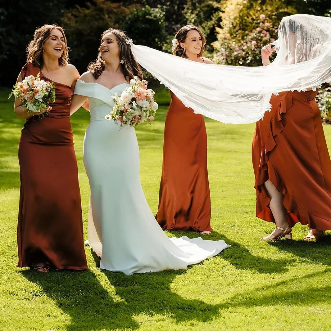 Bridal party outdoors, bride in white gown, bridesmaids in rust-colored dresses, smiling and holding bouquets, with sunlight and greenery in background.