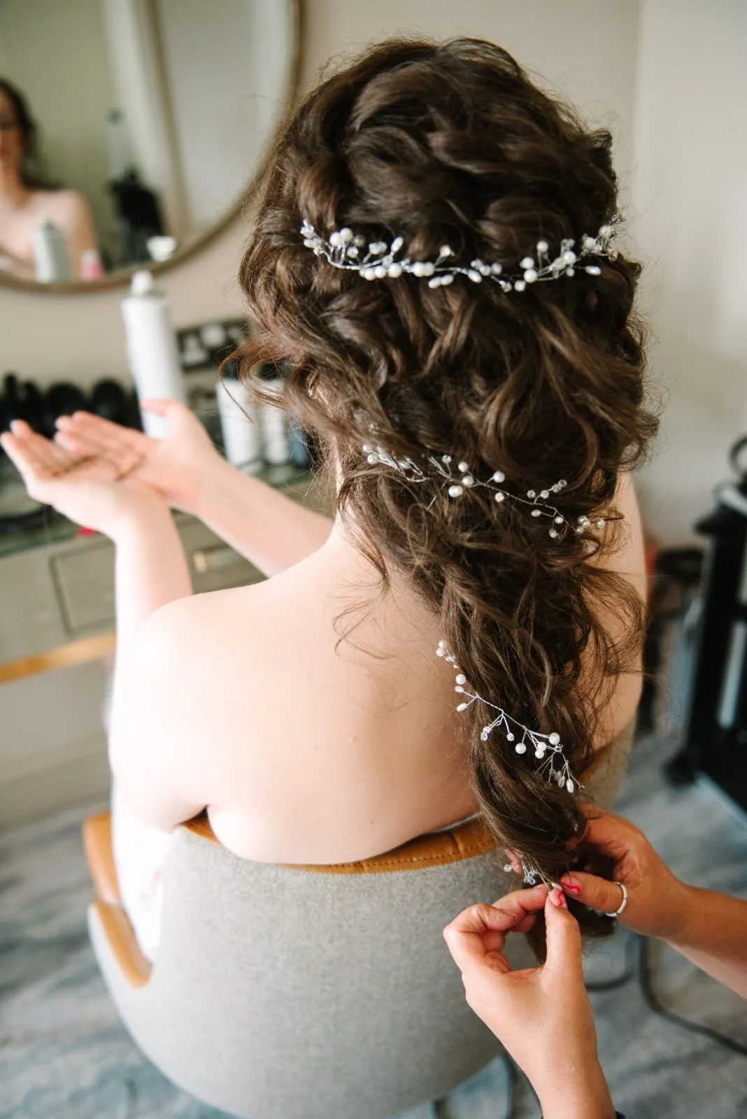 Woman's elaborate curly bridal hairstyle with decorative pearl hairpieces, being done by a stylist in a dressing room.