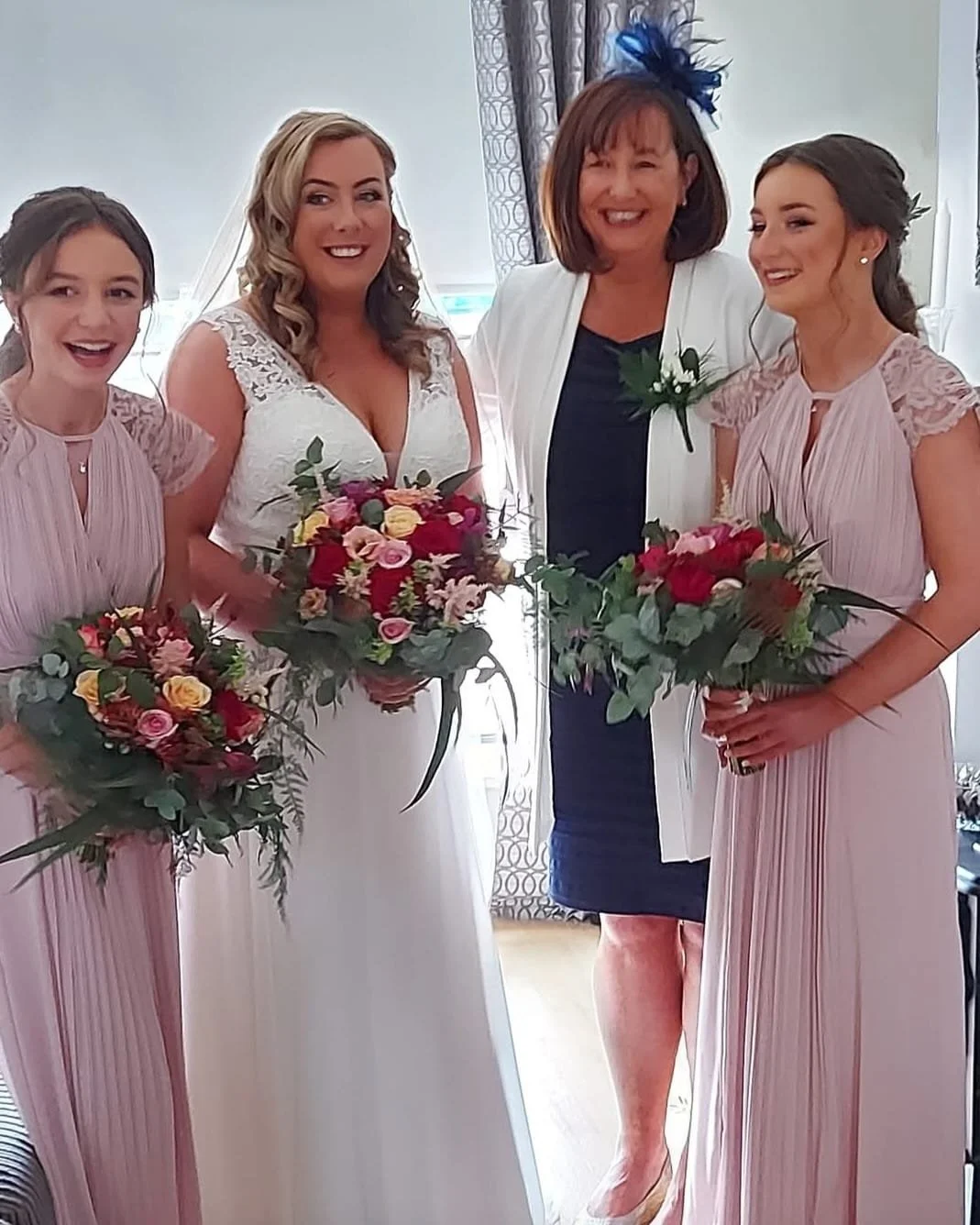 Four women, including a bride in a white wedding dress holding a bouquet, standing with three women in pink dresses and a woman in a black dress with a white blazer and a blue fascinator, smiling inside a room with curtains.