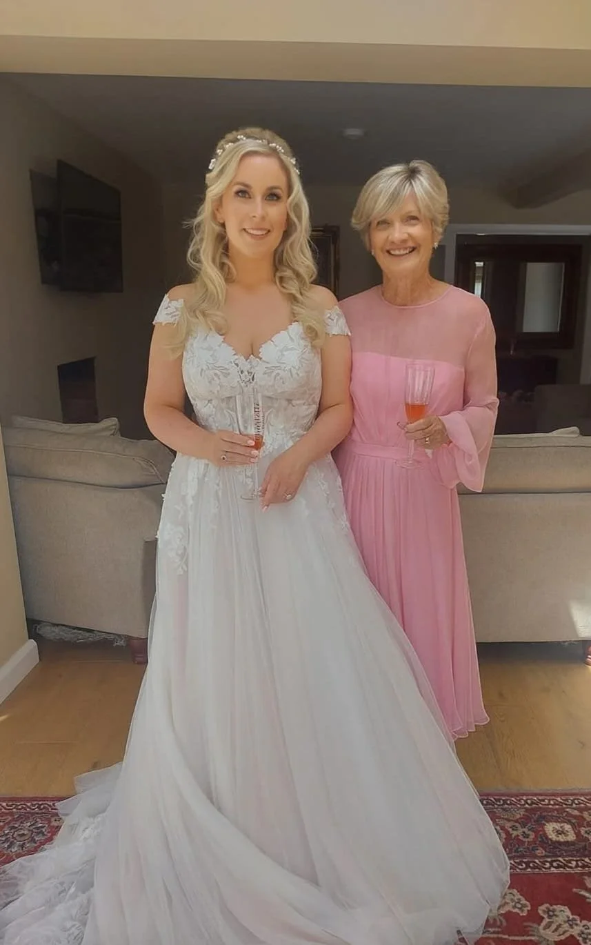 A bride and an older woman, possibly her mother, standing together indoors holding glasses of champagne, both smiling. The bride is wearing a white wedding dress with lace details and a tiara, and the older woman is dressed in a pink gown.