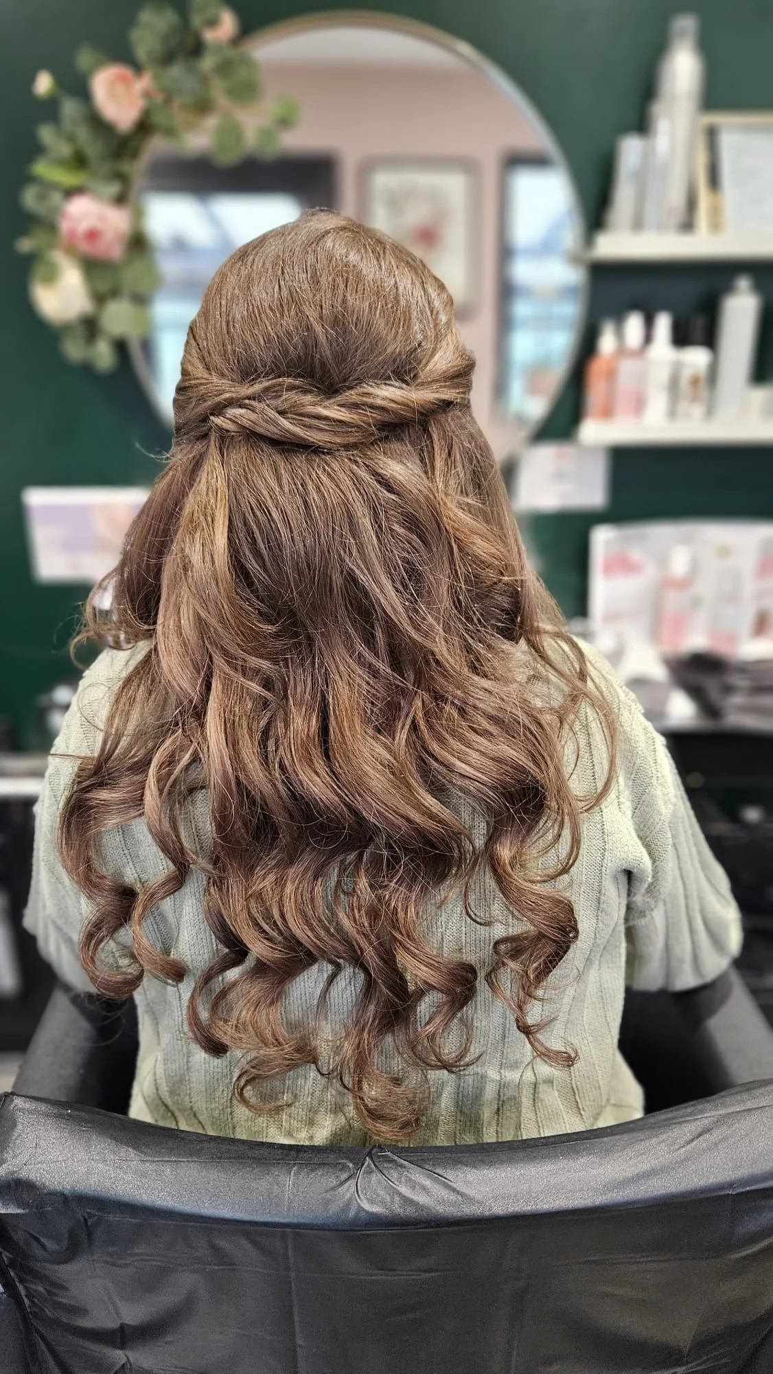 Back of a woman with light brown curly hair sitting in a salon chair, with half of her hair styled into a twisted crown braid and the rest left in loose curls.
