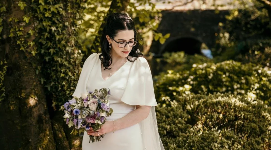 A woman in a white dress holding a lavender and pink bouquet, standing outdoors among greenery with sunlight filtering through the trees.