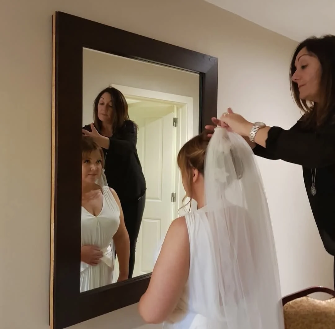 A woman helping a bride with her veil in front of a mirror, with a young girl standing nearby watching.