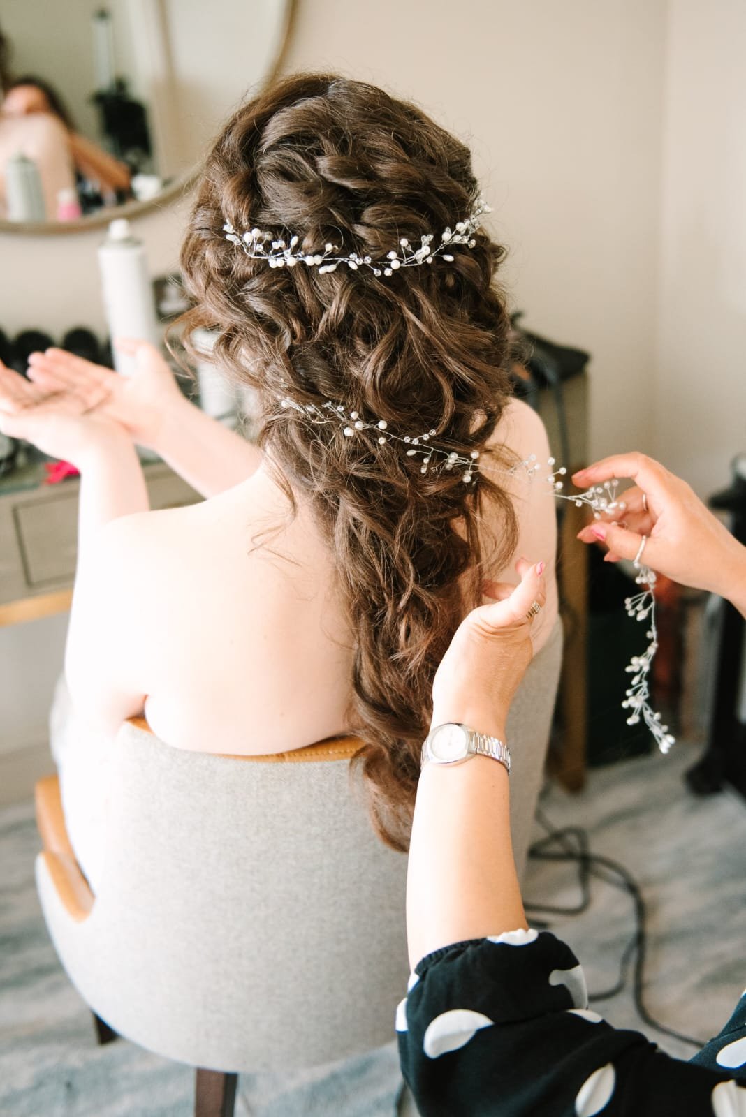A woman with long, curly brown hair is getting her hair styled with decorative pearl and bead hair accessories. Someone is placing a pearl and bead headpiece into her hair, which is being styled in loose curls. The woman is sitting on a chair, facing away from the mirror in a room with a vanity and various beauty products.