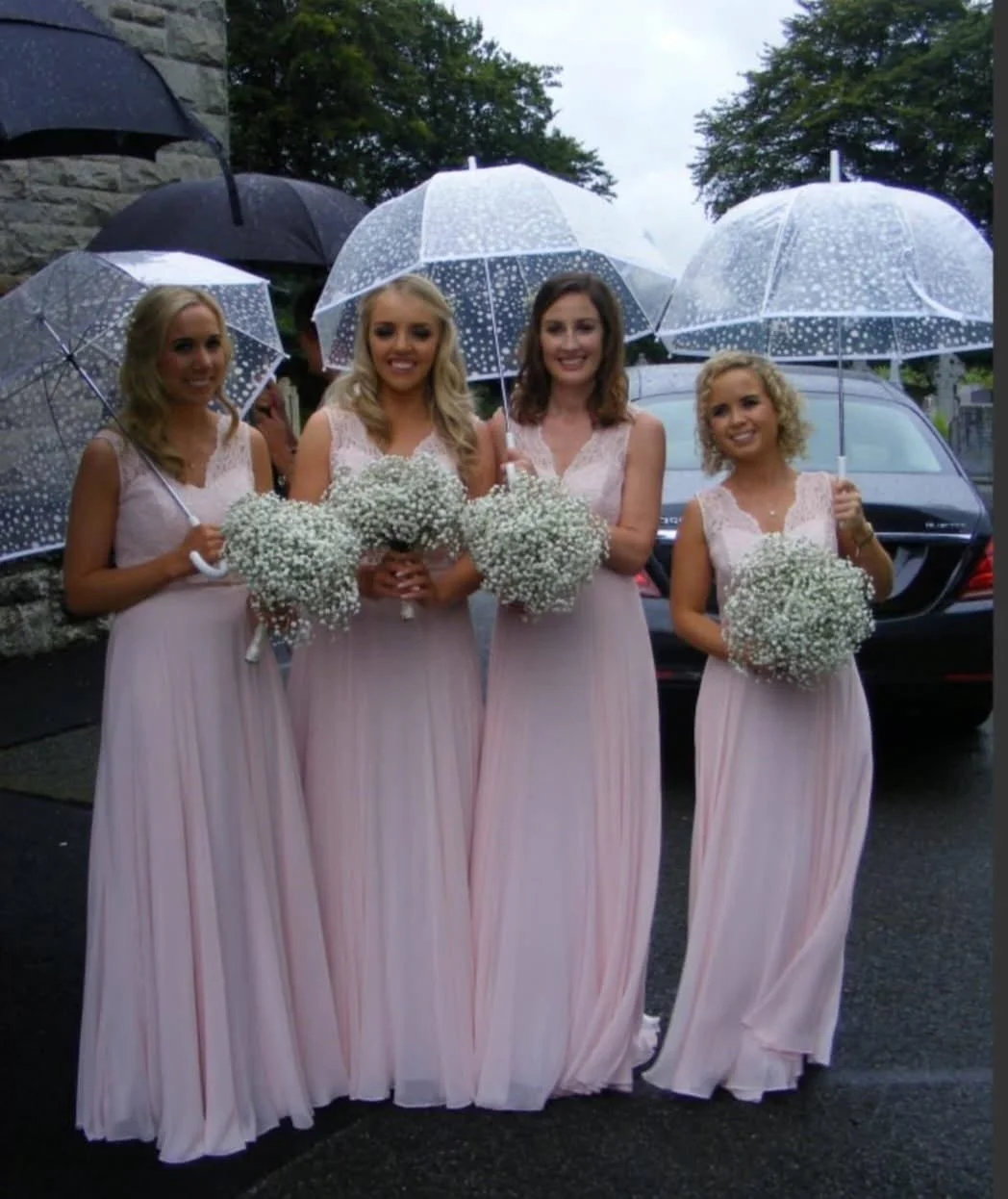 Four women in pastel pink dresses holding bouquets of baby's breath, standing outdoors on a rainy day with umbrellas.