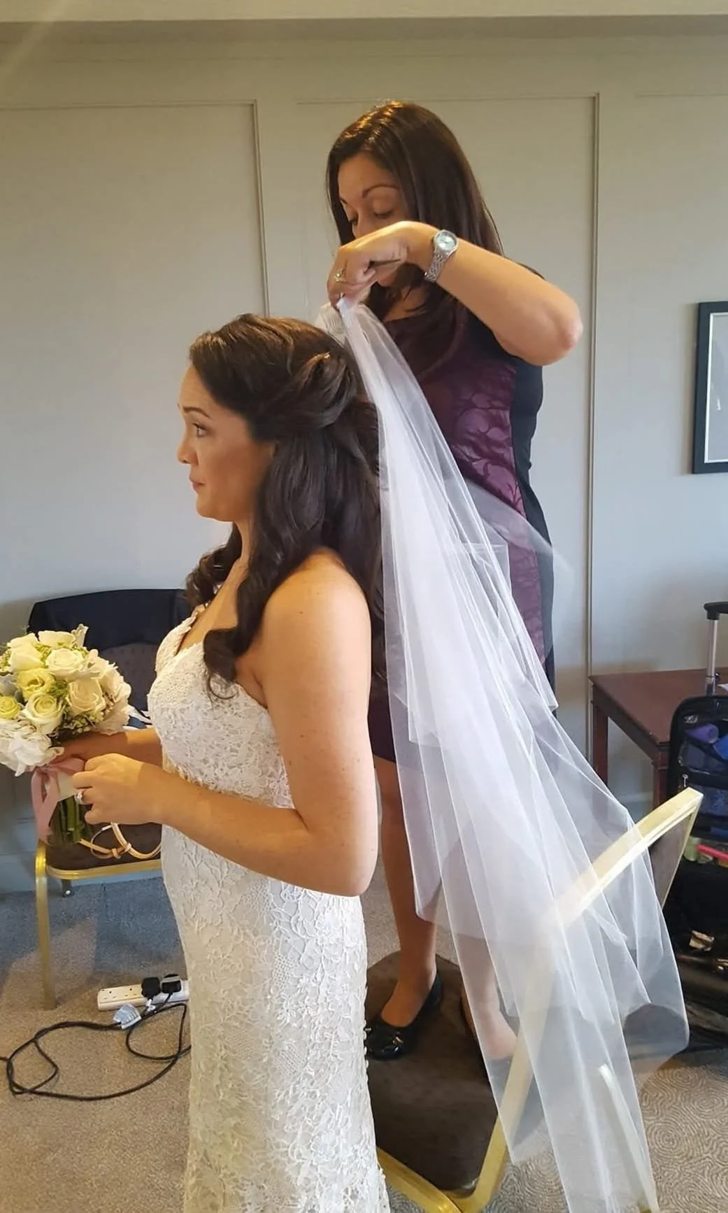 A bride in a white lace wedding dress holding a bouquet of white roses, while a woman adjusts her veil behind her.
