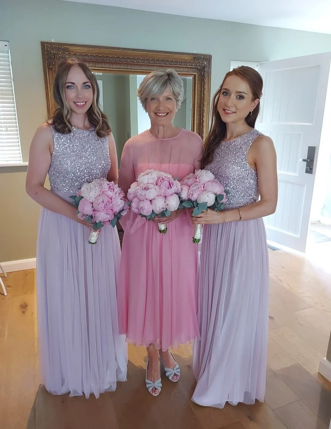 Three women in pink and lavender dresses standing together, holding pink floral bouquets, smiling for the camera. One older woman in the center, flanked by two younger women styled as bridesmaids.