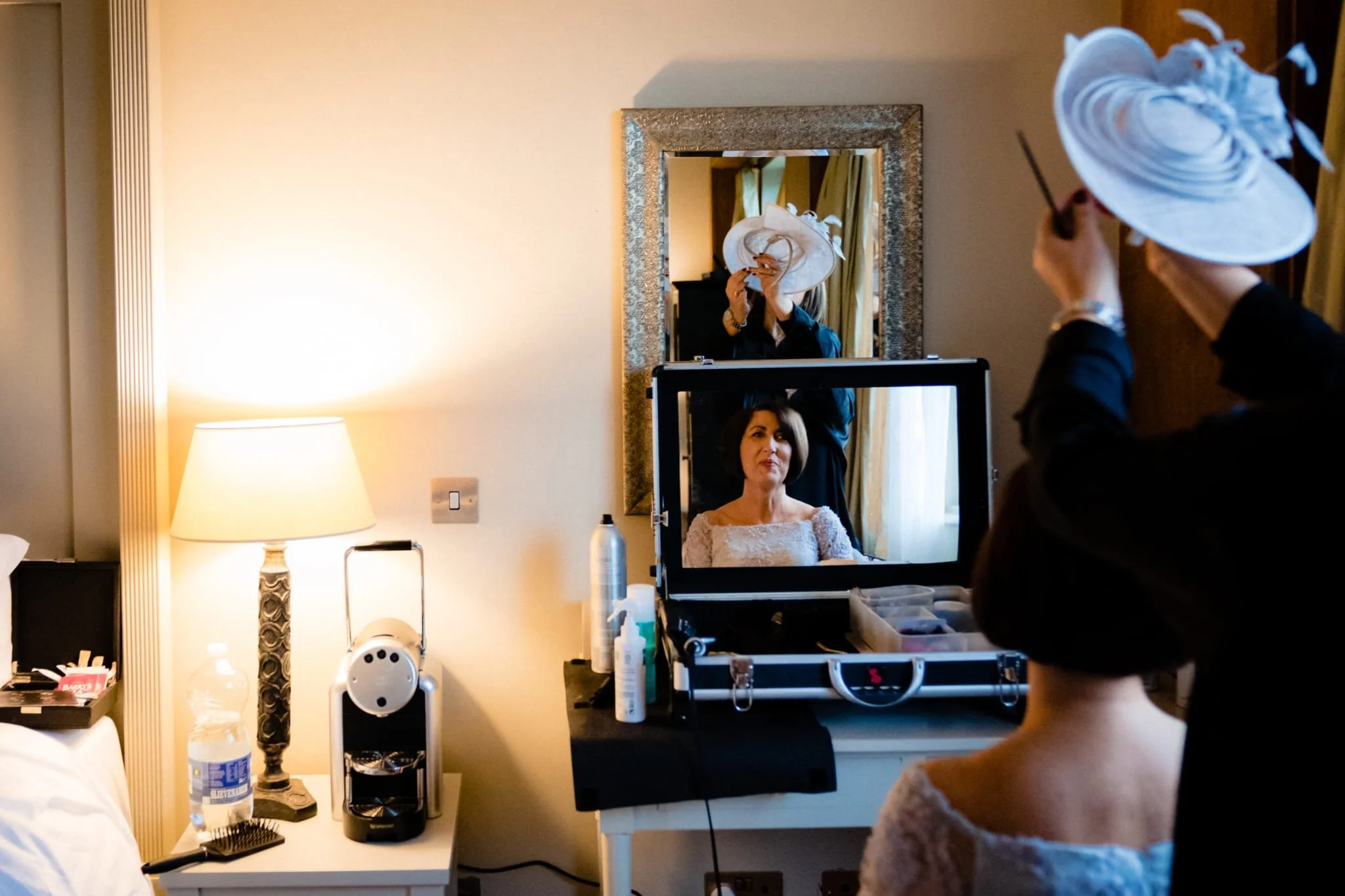 A woman is sitting in front of a mirror while a makeup artist applies makeup and adjusts her hat. The woman's reflection is visible in a TV screen on the dresser. The room has a lamp, water bottle, and beauty products on the side table.