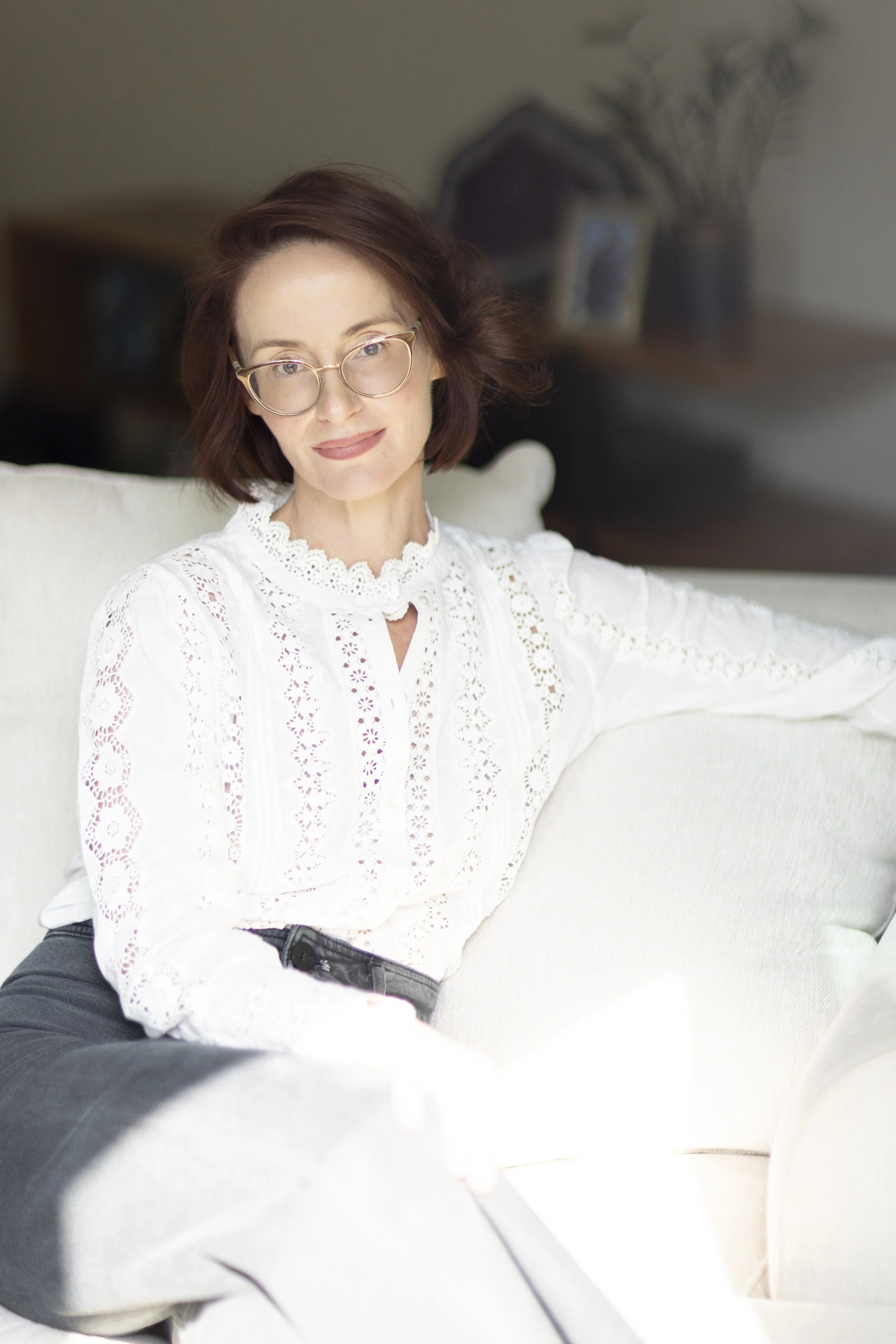 The author of Happy Mr Bond, Charity L Adams, wearing glasses and shoulder-length brown hair with a white lace blouse and black jeans, sitting on a cream-coloured sofa, behind which is a blurred background.