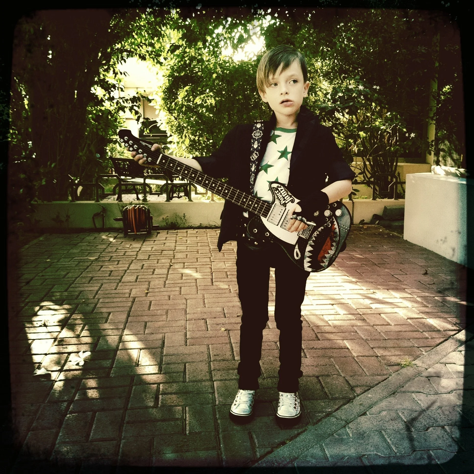 A young boy named Bond is dressed like a rockstar, holding a guitar and wearing a black jacket, star-patterned shirt, skinny black jeans, and white shoes. There are trees and benches in the background, with sunlight filtering through the leaves.