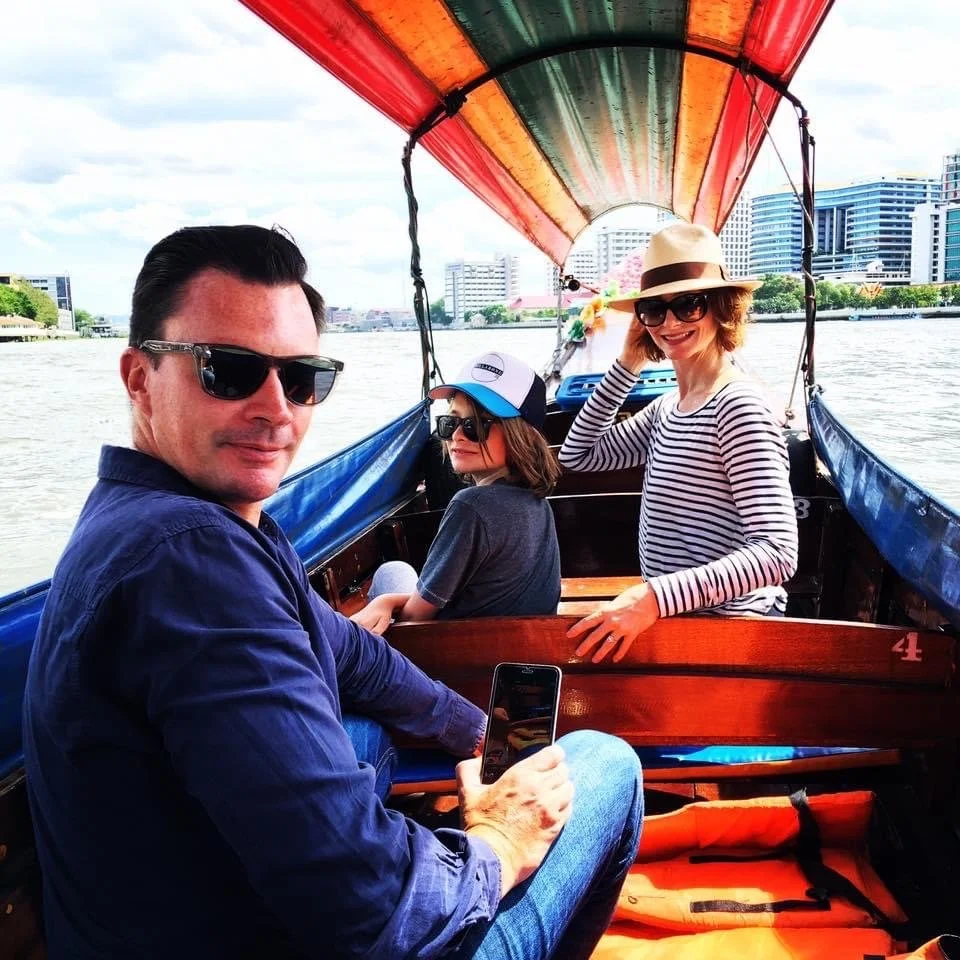 A family photo set within a Bangkok riverboat enveloped by a colourful canopy and glistening buildings on the riverbank. The father is wearing a navy shirt, the son in a grey t-shirt and cap, and the mother in a rattan panama hat and striped top.