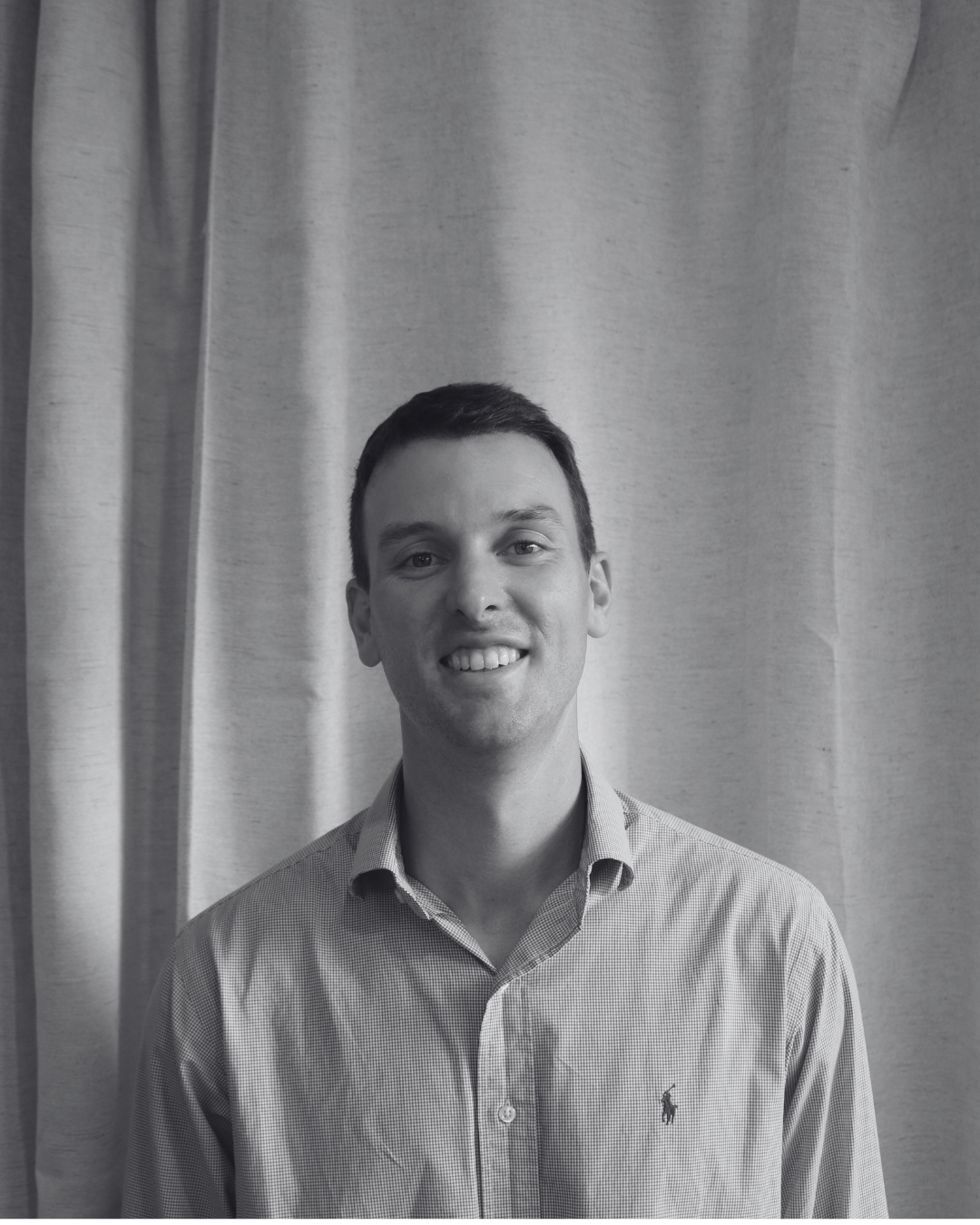 A black and white photo of a young man smiling, standing in front of a curtain.