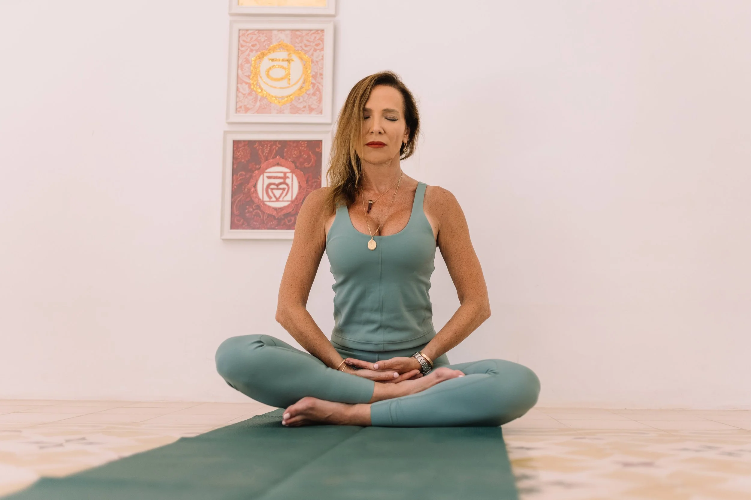 A woman practicing yoga indoors on a blue mat near an open window with a scenic mountain view, smiling and sitting cross-legged.