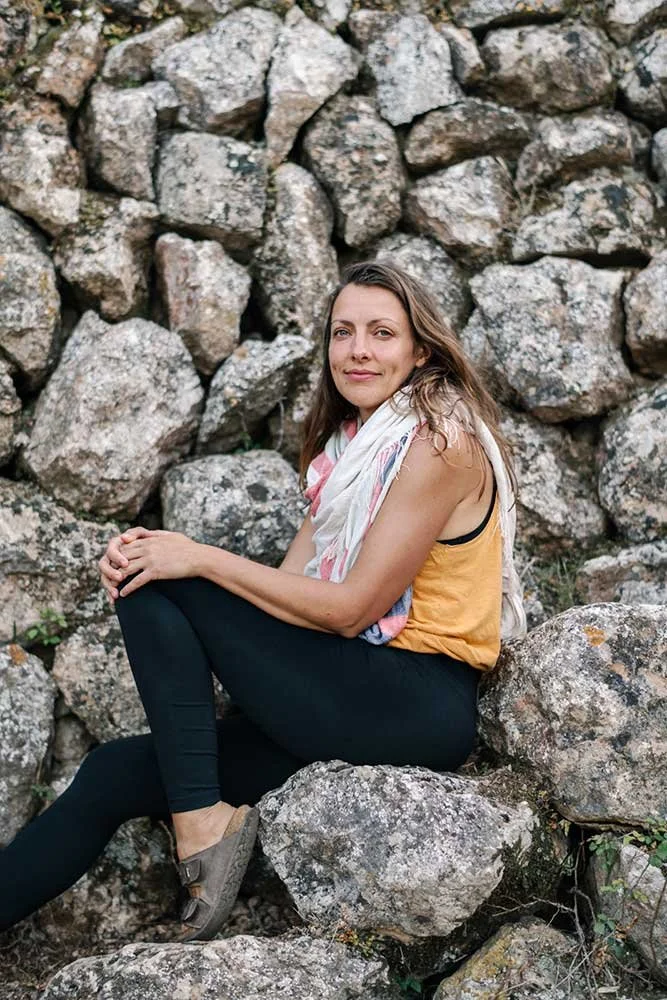 A woman practicing yoga outdoors during the day, standing on one leg in a tree pose under a large leafy tree.