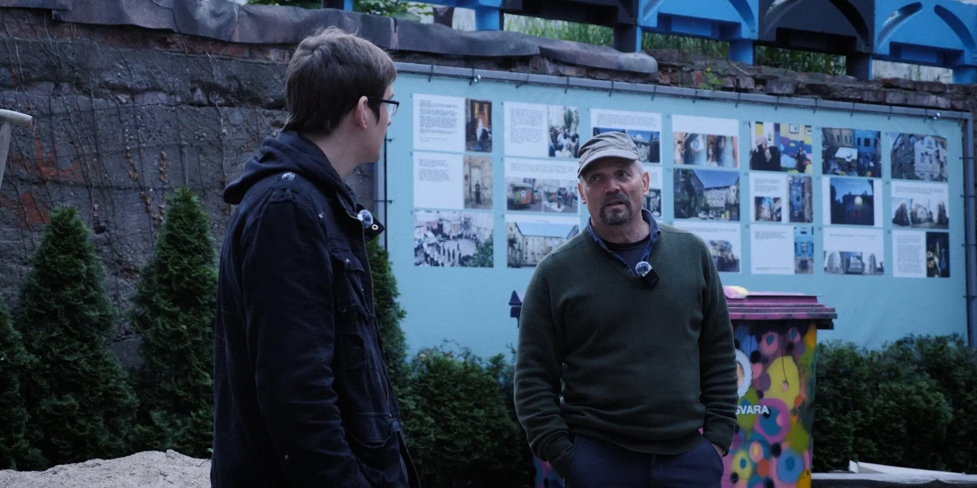 Two men, one with glasses and a dark jacket, and the other with a cap and green sweater, are having a conversation outdoors near display boards with photos and text. There are small bushes and a painted utility box behind them.
