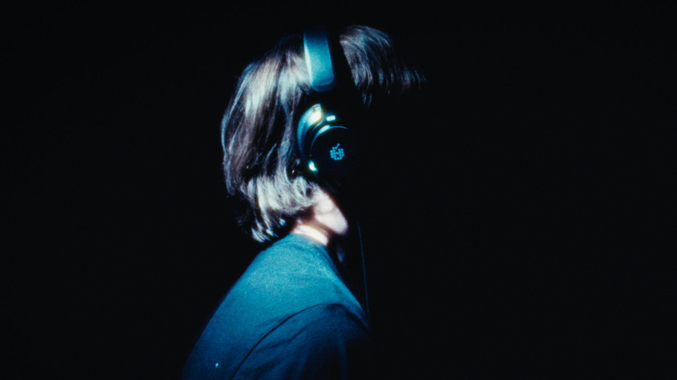 Side view of a person with shoulder-length hair wearing headphones against a dark background.