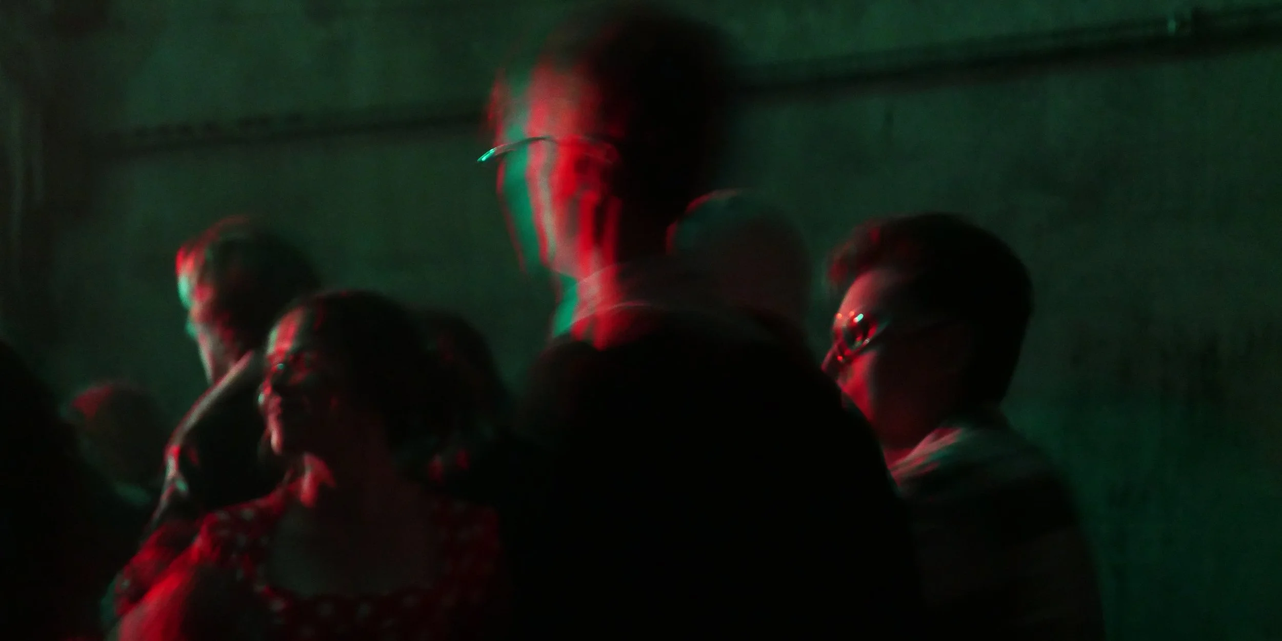 A group of people standing in a dark, green-tinted environment, with some red and green lighting effects. The photo is blurry, and the individuals are facing different directions.