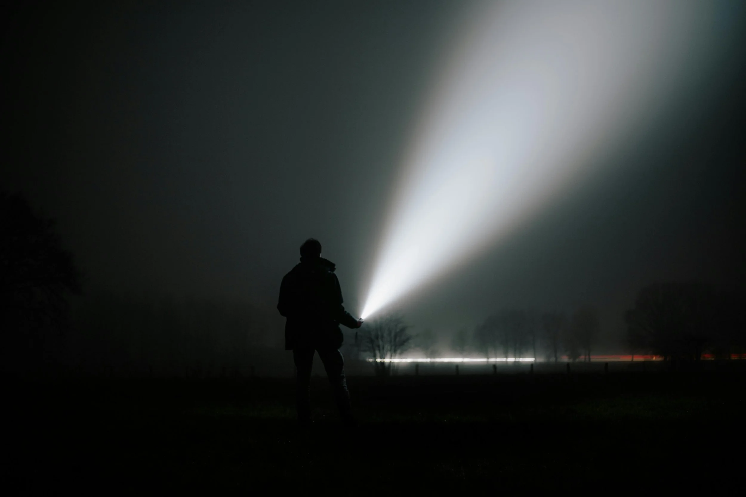 Person holding a flashlight in a dark outdoor setting at night, with the beam illuminating into the sky.