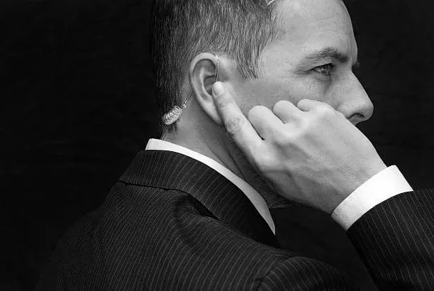 A man in a suit touching his ear with his hand, viewed from the side against a black background.