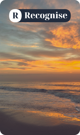 A scenic view of a sunset over the ocean with a lighthouse on a distant cliff, a beach with rocks in the foreground, and a sky painted in shades of purple, pink, and orange.