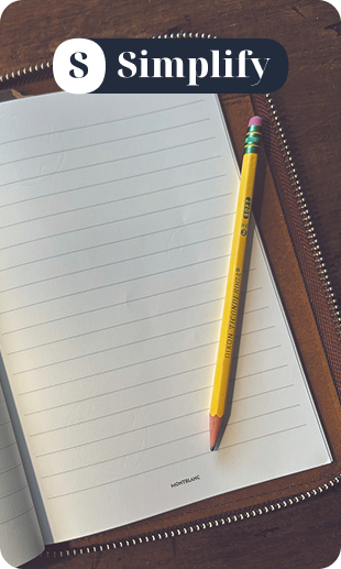 A desk with a spiral notepad, two red pencils, a brown paper card, a concrete pot with a succulent plant, a small black and white object, and a wooden box with mechanical parts.