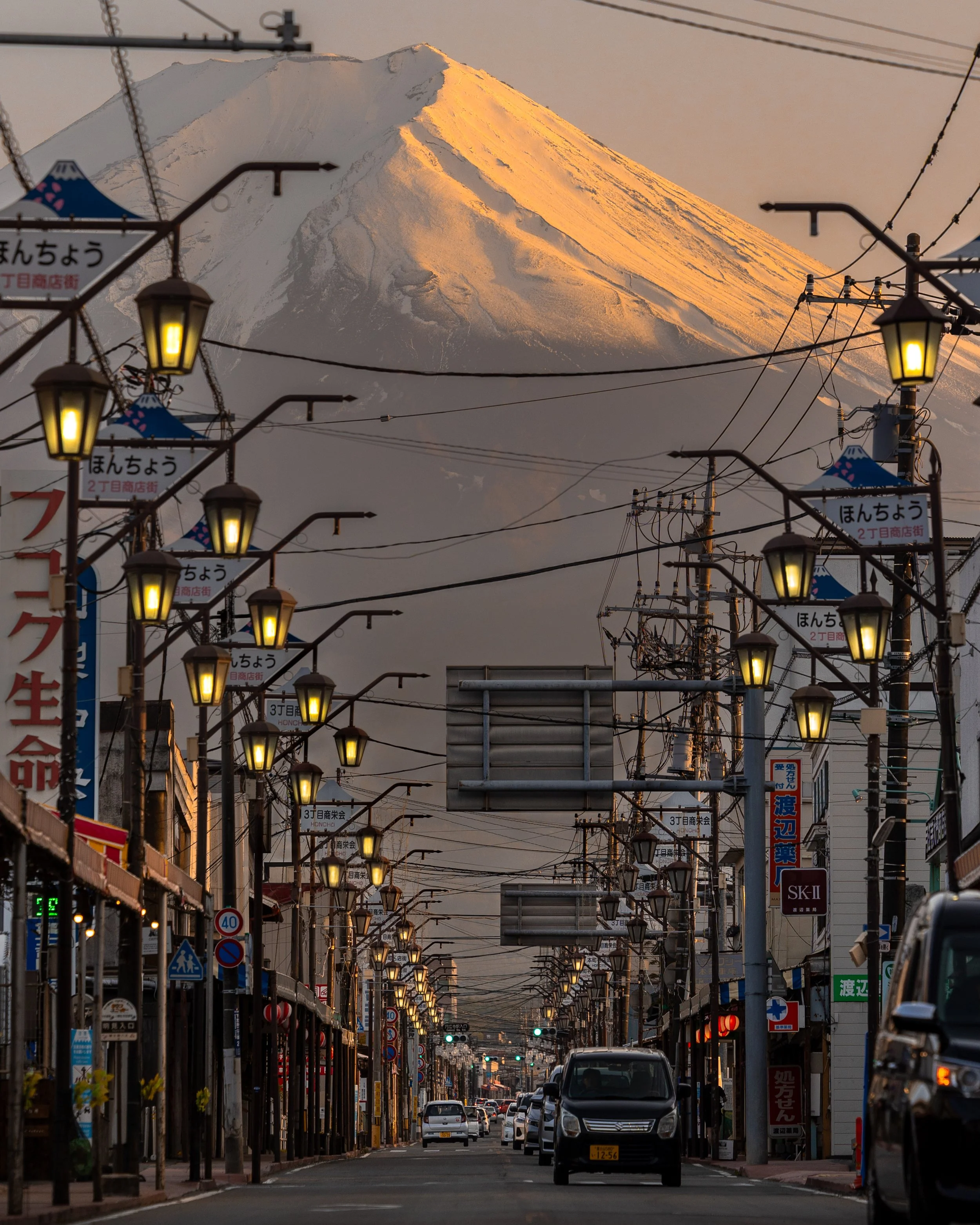 Street view with cars and shops, leading towards a snowy mountain peak, illuminated by sunlight, in the background.