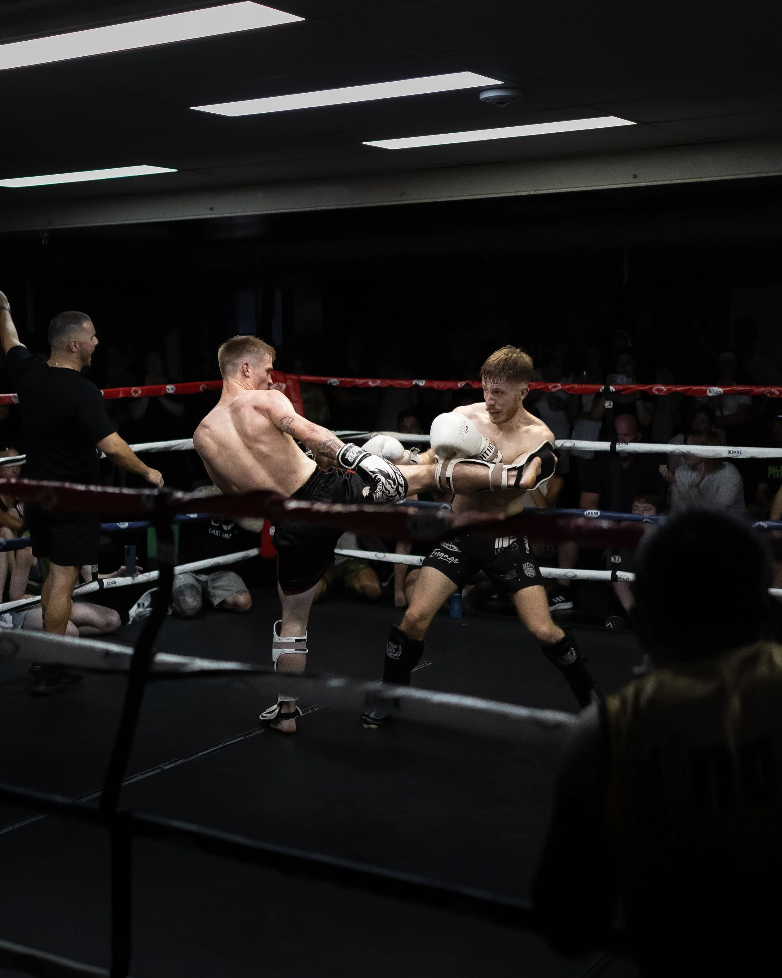 Two men boxing in a ring during a match, wearing shorts and boxing gloves, with spectators in the background.