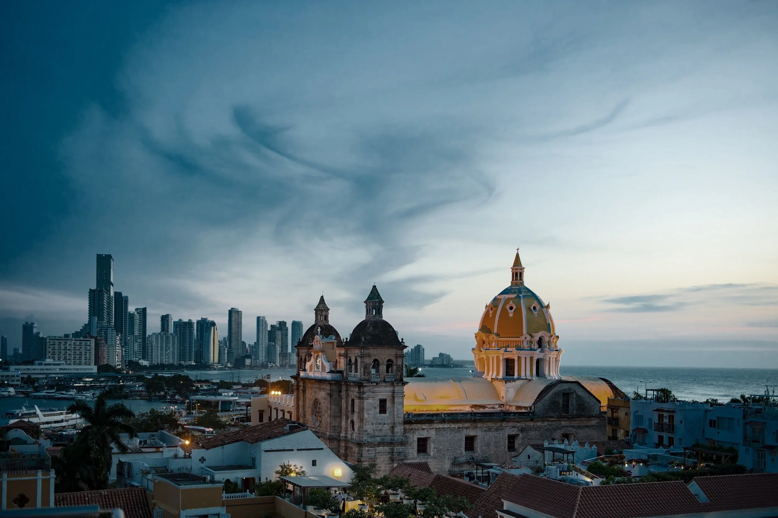 Stadtansicht mit historischem Kirchengebäude am Meer bei Sonnenuntergang, moderne Hochhäuser im Hintergrund.