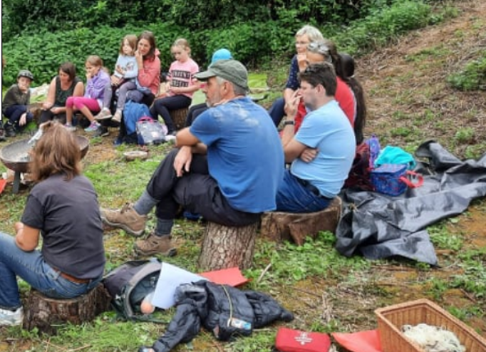 Group of people sitting outdoors on logs and the ground in a grassy area with trees, participating in a workshop or storytelling session.