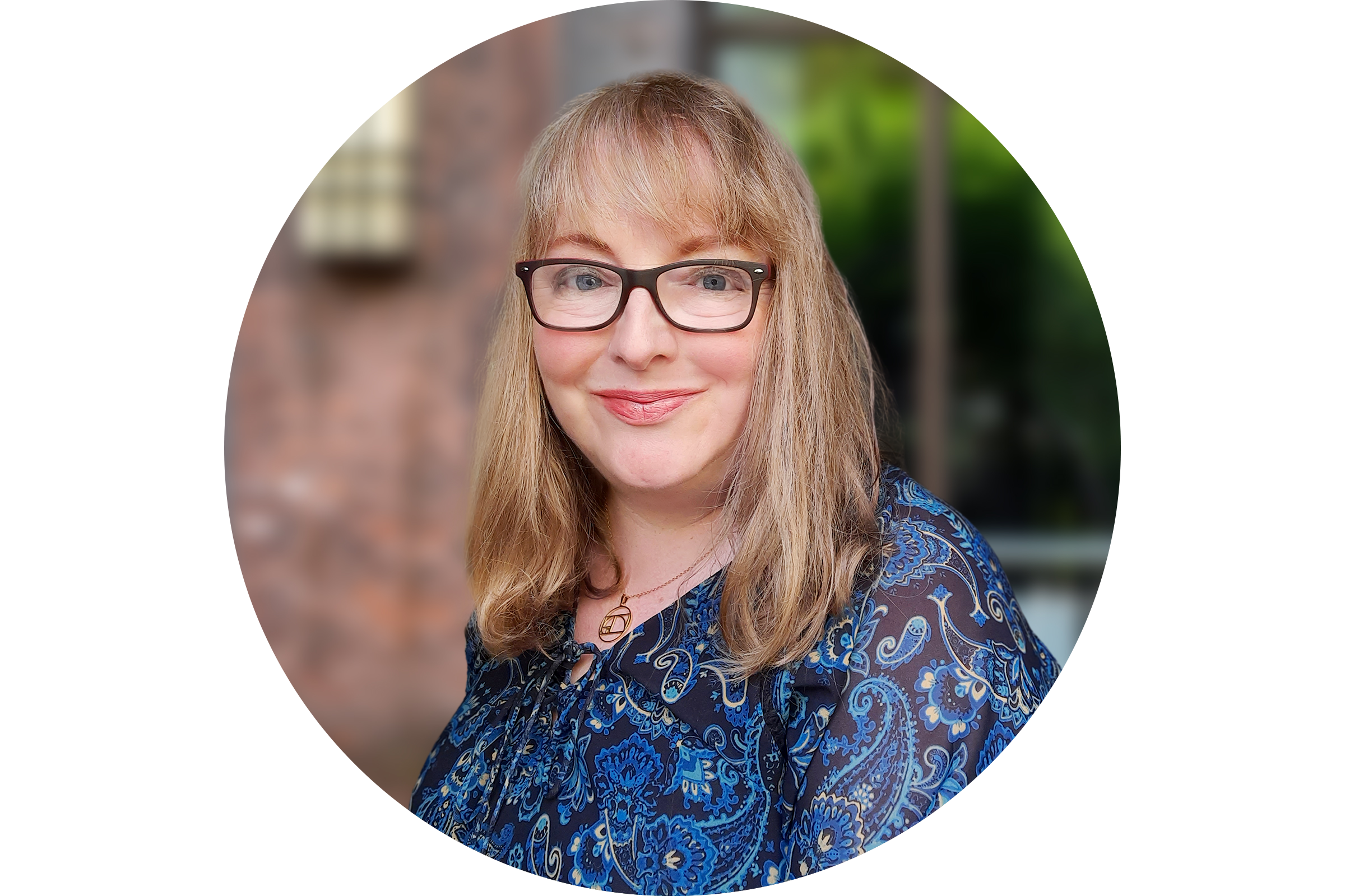 Portrait of a woman with shoulder-length blonde hair, wearing glasses and a blue paisley blouse, smiling outdoors with a blurred background.