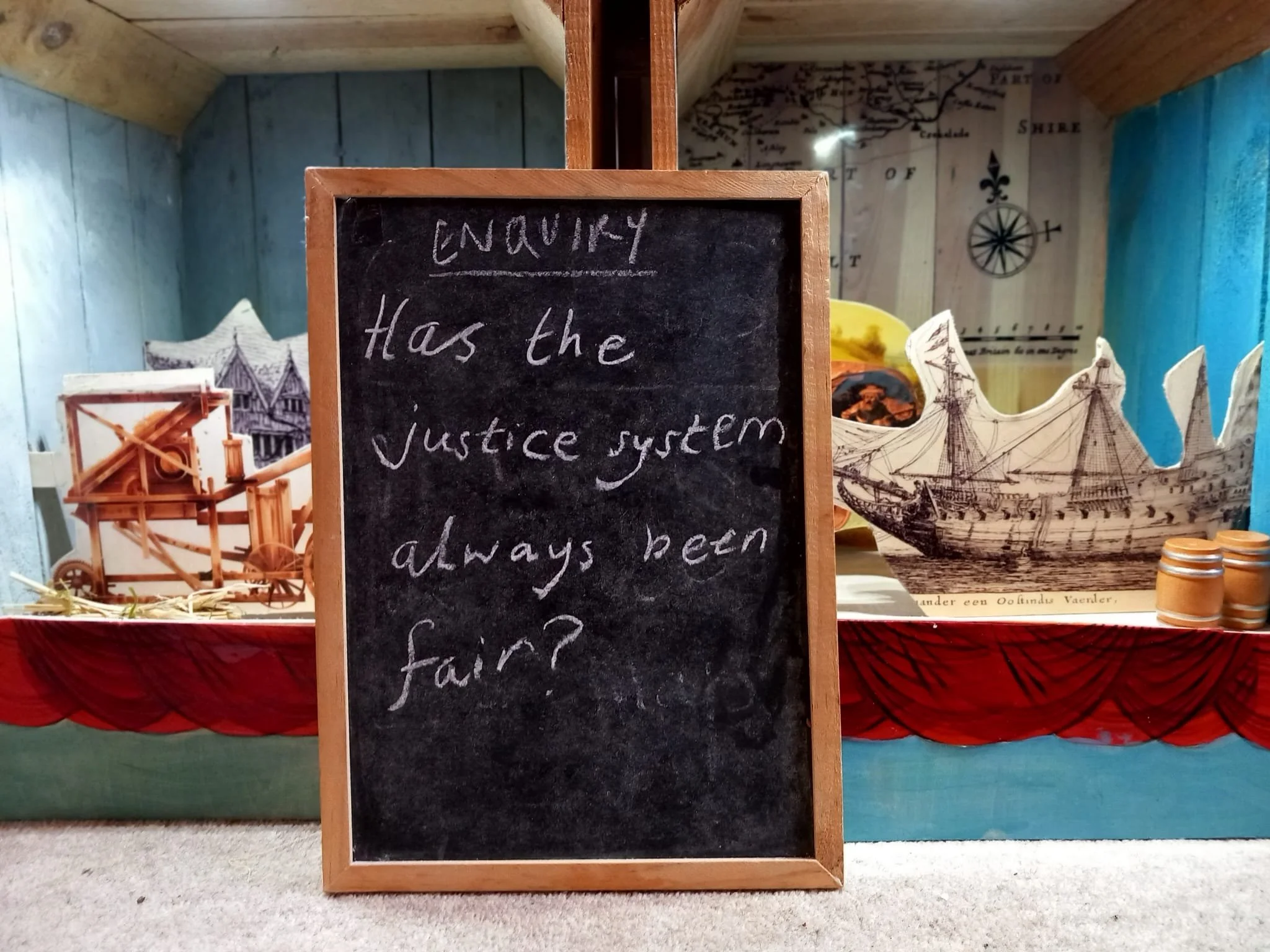 A black chalkboard with the handwritten question, 'Does the justice system always been fair?' in white chalk, positioned in front of a display with a model ship, barrels, and a map-themed background.