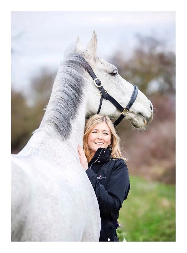 A woman smiling and hugging a large white horse outside.