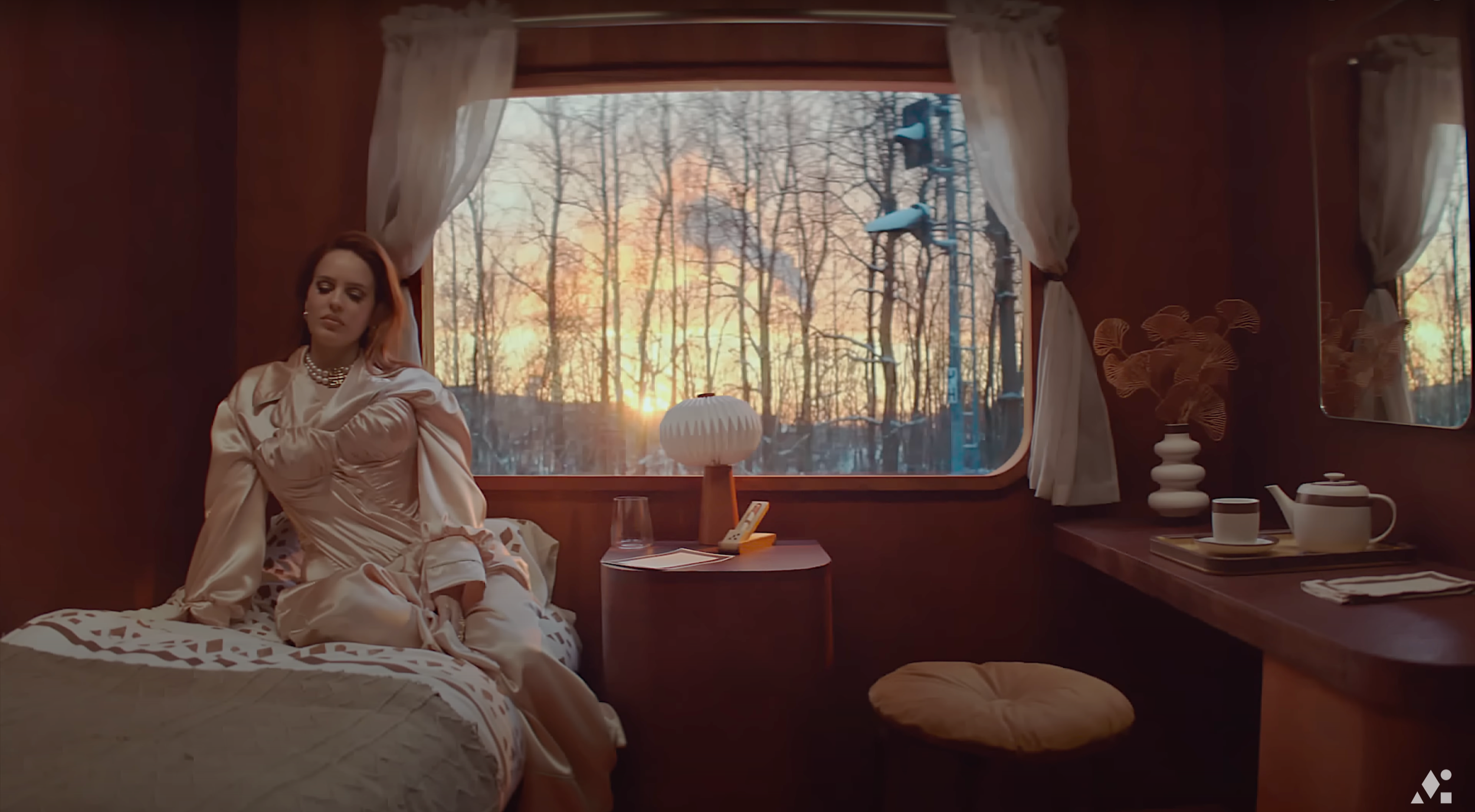A woman with brown hair sitting on a bed inside a cozy wood-paneled room. She is dressed in satin pajamas and jewelry, looking contemplative. Behind her, a large window shows a winter scene with bare trees and a sunset. The room has a bedside table w