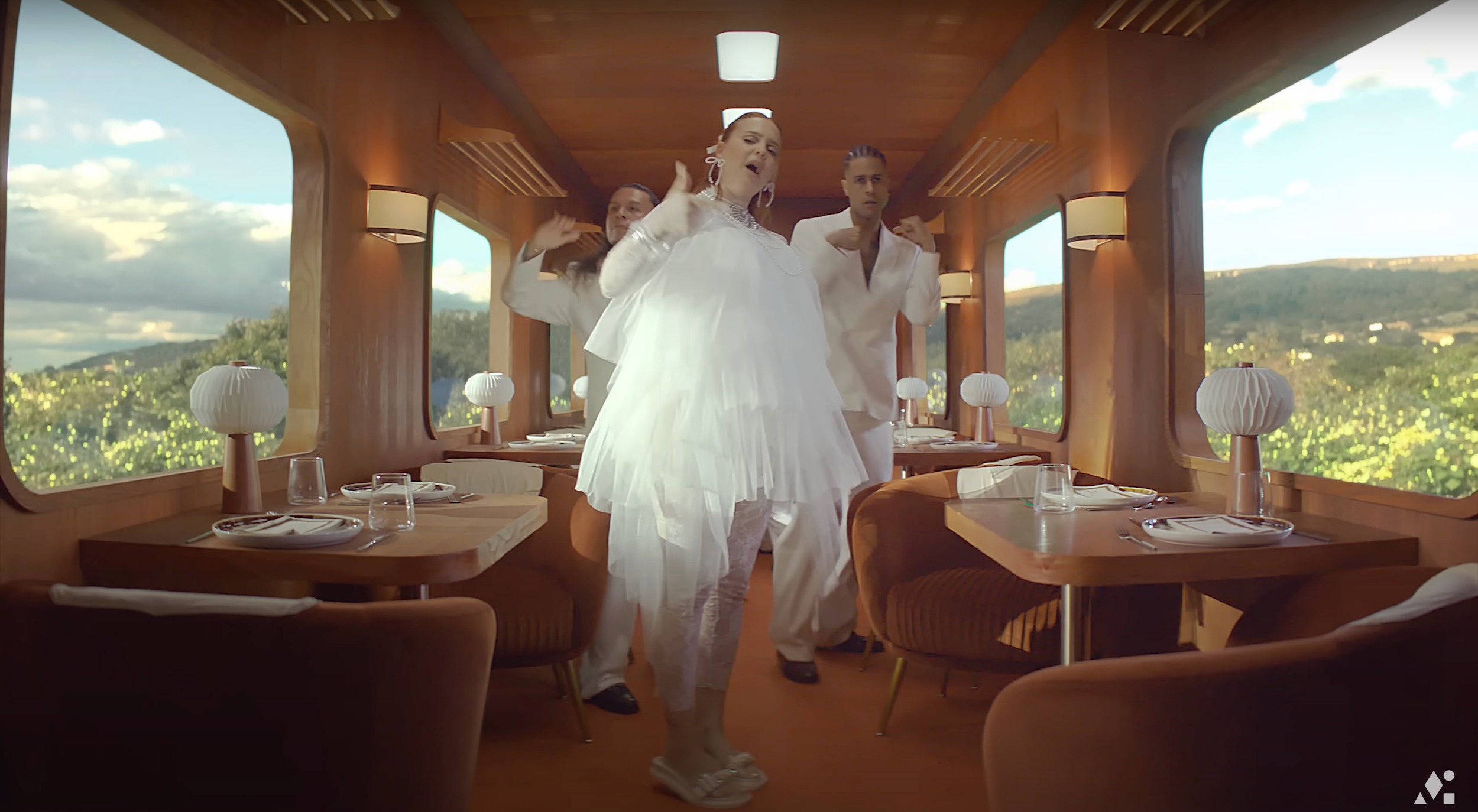 Three women in white clothing dancing and singing inside a dining car with large windows showing a scenic landscape outside.