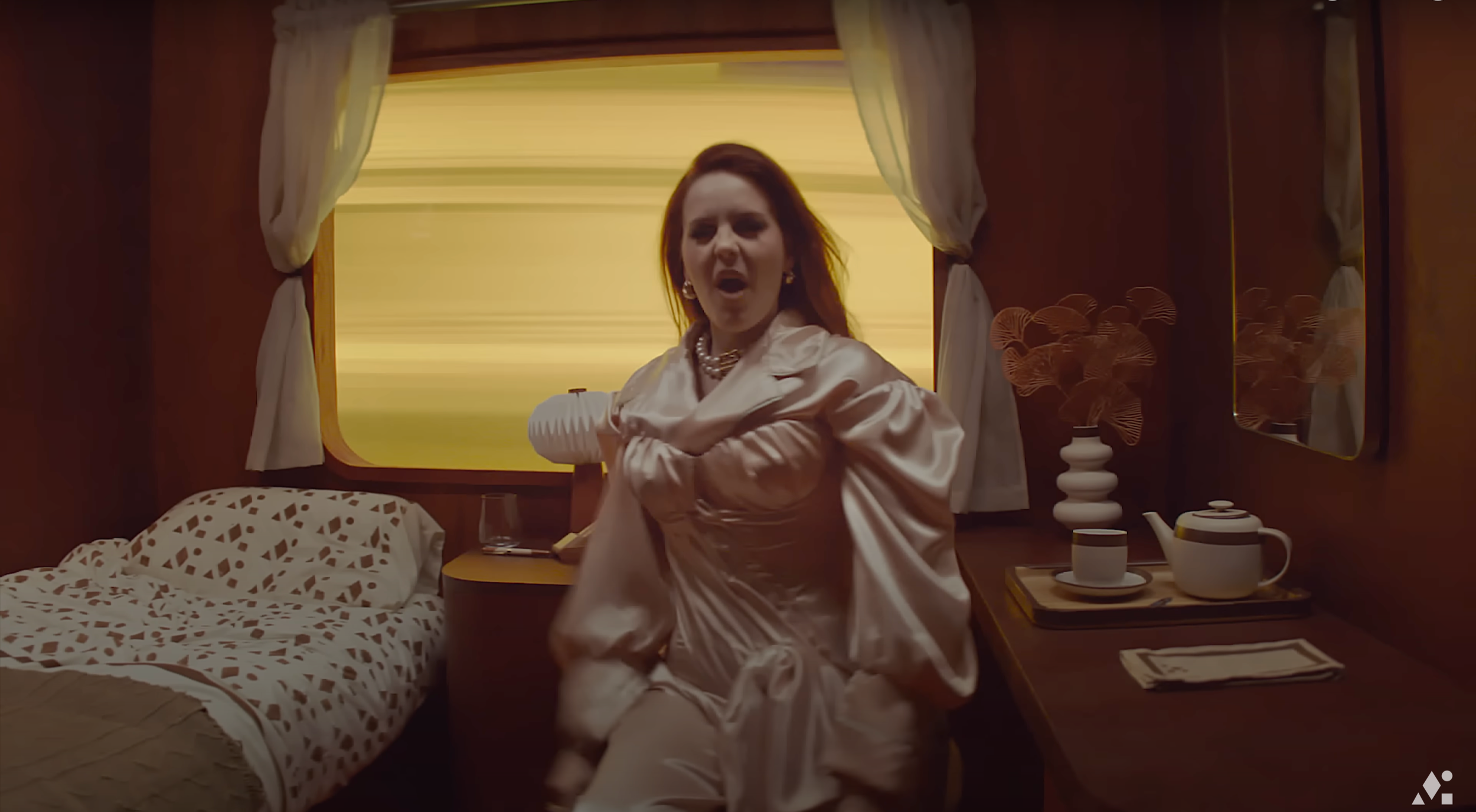 A woman with red hair, wearing a shiny cream-colored satin dress and pearl necklace, is inside a vintage train cabin. She appears to be angry or yelling, with her hand clutching her chest. The cabin has a bed with geometric bedding, a small bedside t