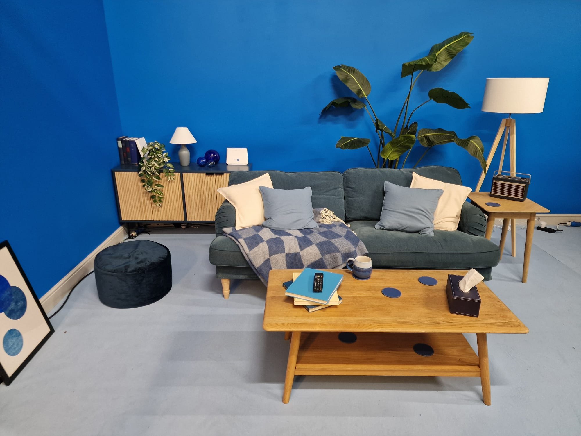 Living room with a dark green sofa, pillows, a blanket, a wooden coffee table with books and a mug, a side table with a lamp, a large potted plant, a blue painted wall, a cabinet with books and decorations, and a framed art piece on the floor.