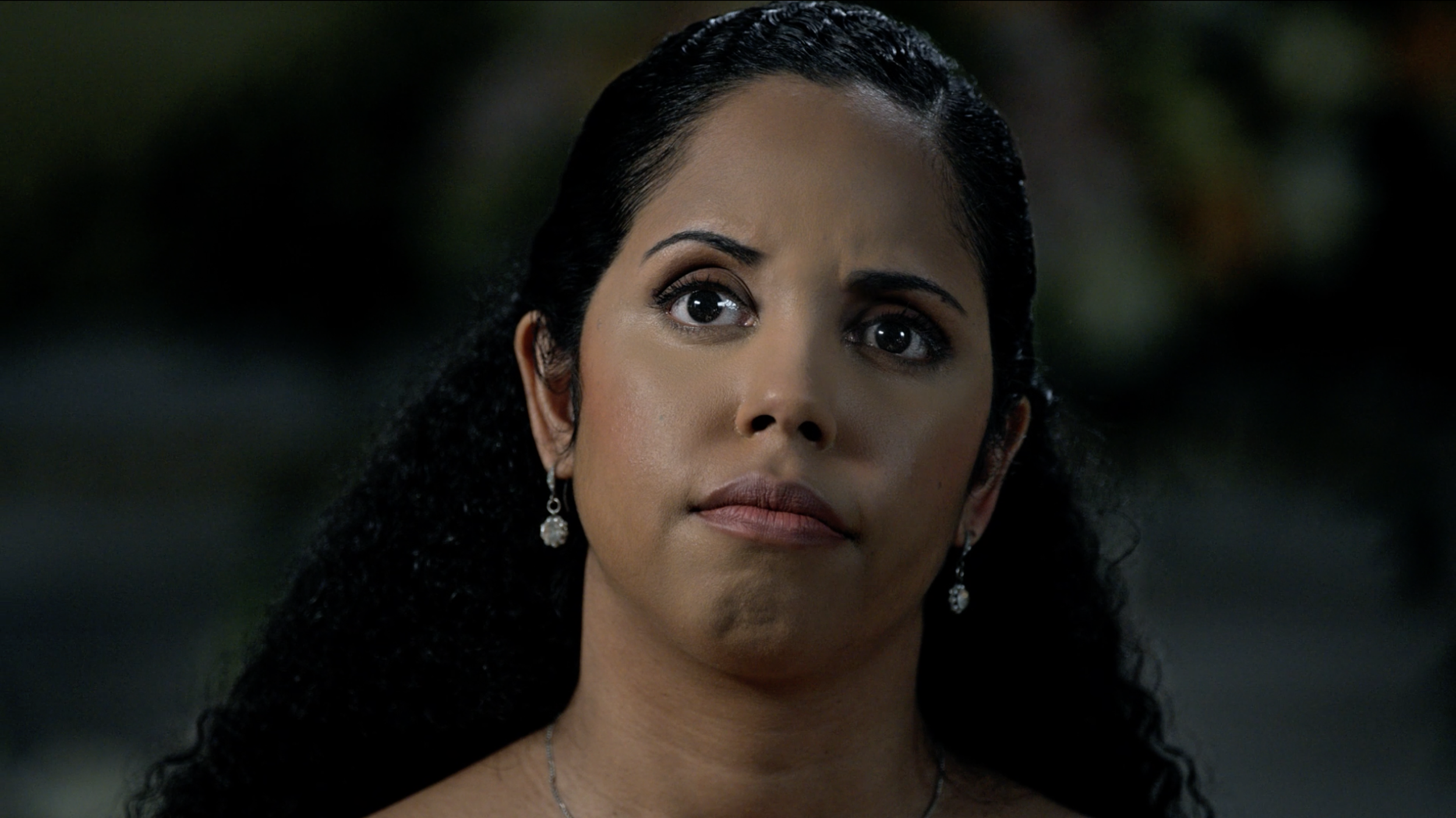 Close-up of a woman with dark, curly hair wearing earrings and a necklace, with a serious or contemplative expression on her face, outdoors at night.
