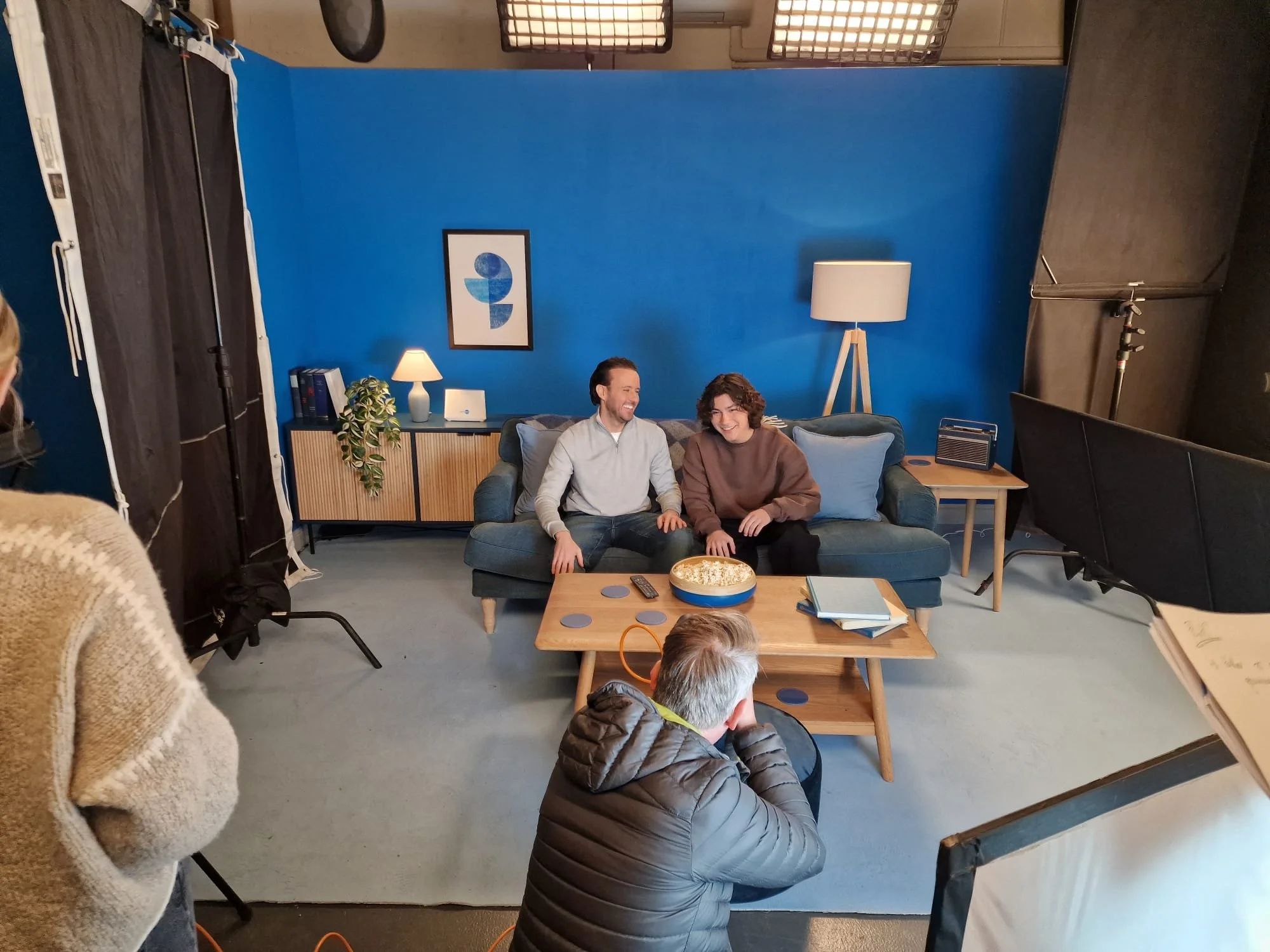 Two people sit on a blue sofa, laughing, during a filmed interview in a studio with a blue wall. A person in a gray jacket is seated on a lower chair with a camera directed at them. Studio equipment such as lights and a partition are visible.
