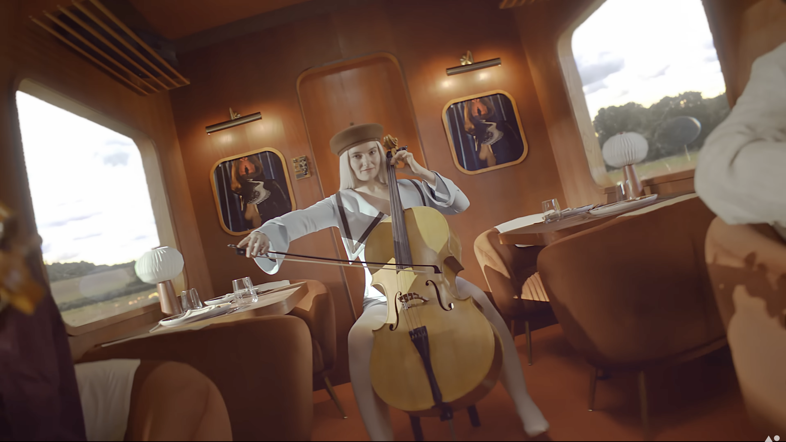 A woman wearing a beige beret and a white coat playing a cello with a bow inside a train carriage with wooden interior and tables set for dining.