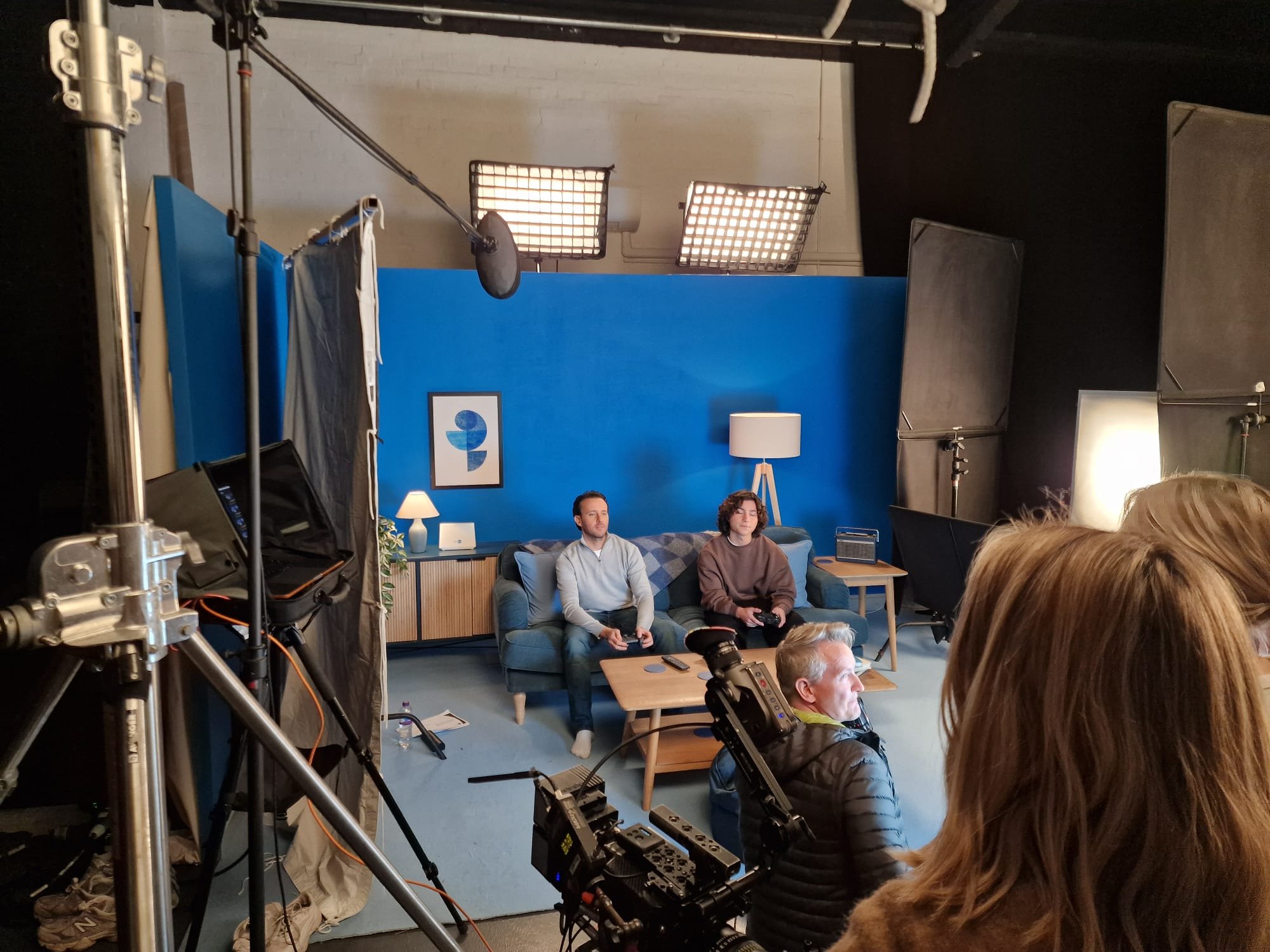 Two people sit on a blue couch in a TV studio with a blue wall behind them. They hold game controllers and are in front of a wooden coffee table. Studio lighting equipment and cameras are visible in the foreground, and a small audience watches from t