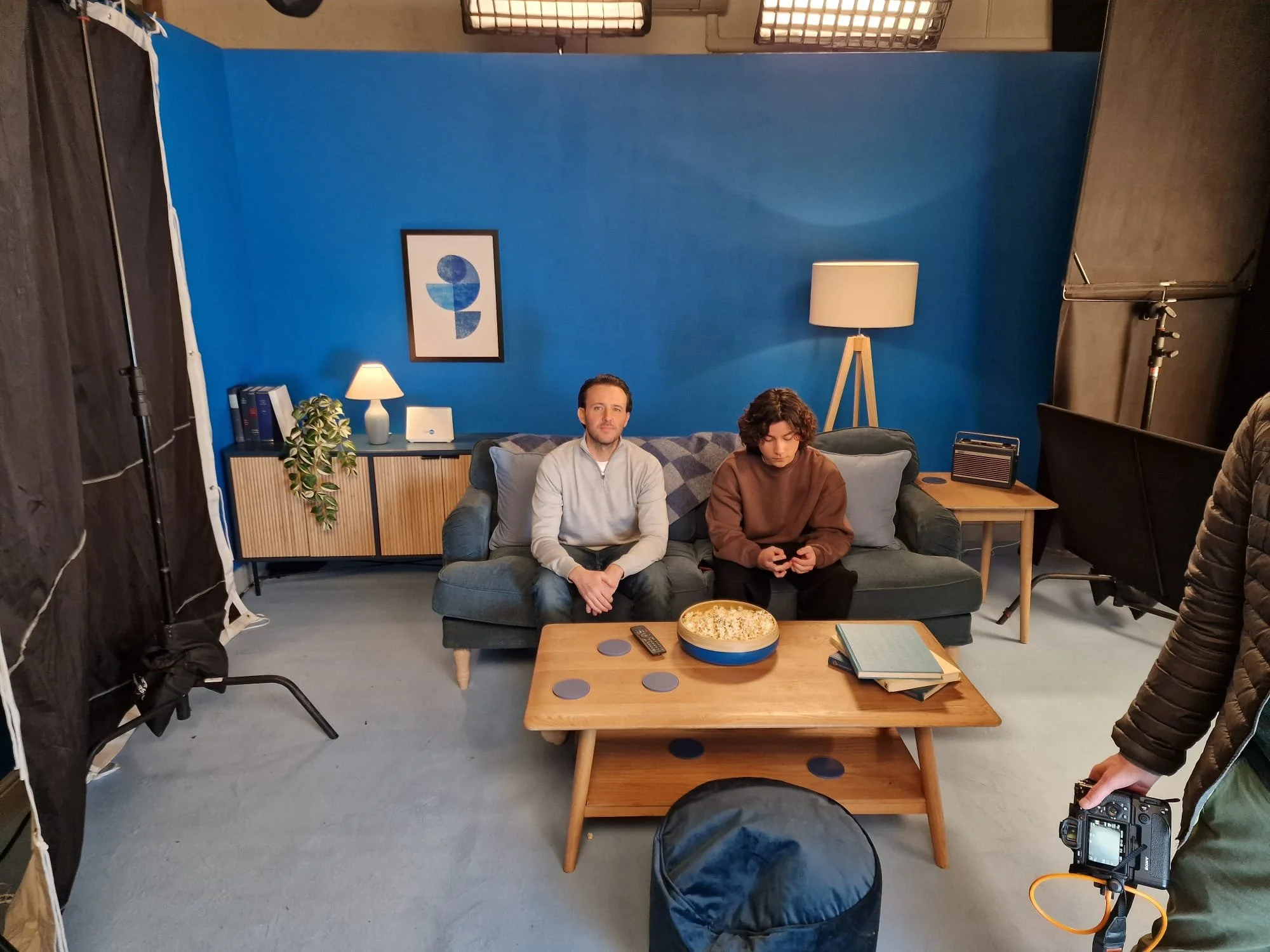 Two people sitting on a blue couch in a studio setup, with a blue wall behind them and a framed abstract art piece. One person looks at the camera, the other looks down at their phone. A coffee table with food and books is in front of them. Studio eq