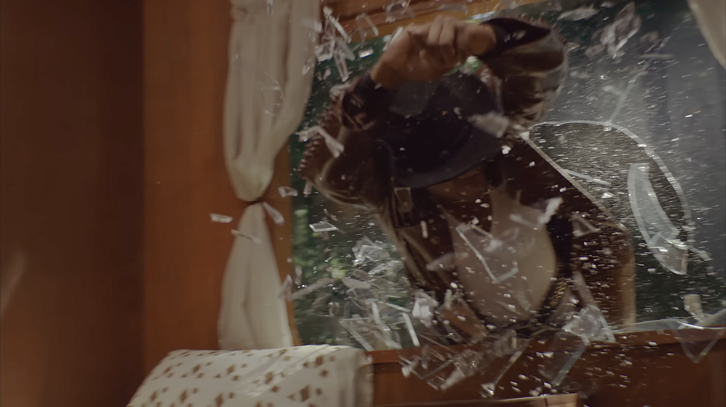 A person in dark clothing breaking through a glass window, shattering glass pieces around and on them, with furniture and a window in the background.