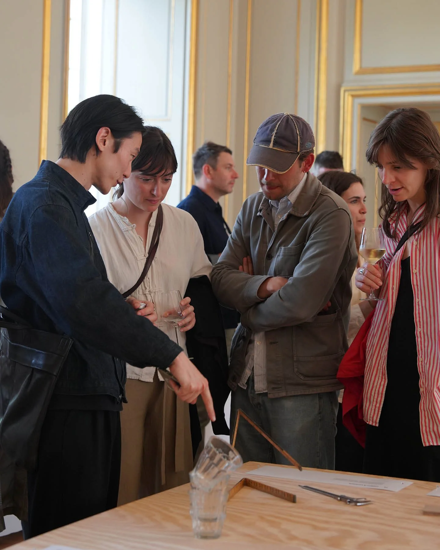 A group of five adults, four women and one man, gathered around a table with glasses of wine, attentively looking at and discussing a small object or demonstration during a social event in a decorated room.