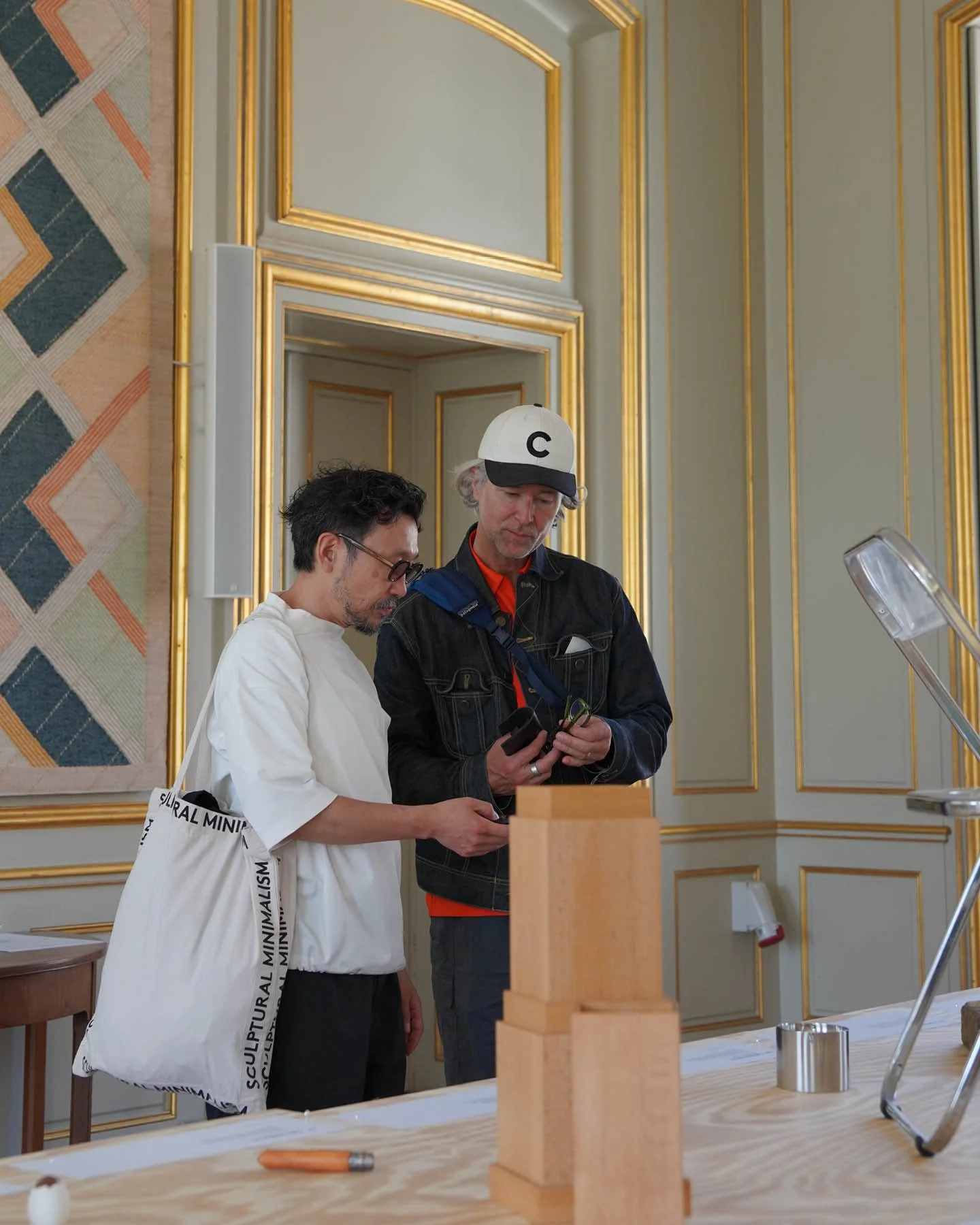 Two men are standing at a table indoors, looking at a device. One man is wearing a white t-shirt and glasses, with a white bag over his shoulder, and the other is wearing a black jacket, red shirt, and a white cap with a black letter 'C'. There is a
