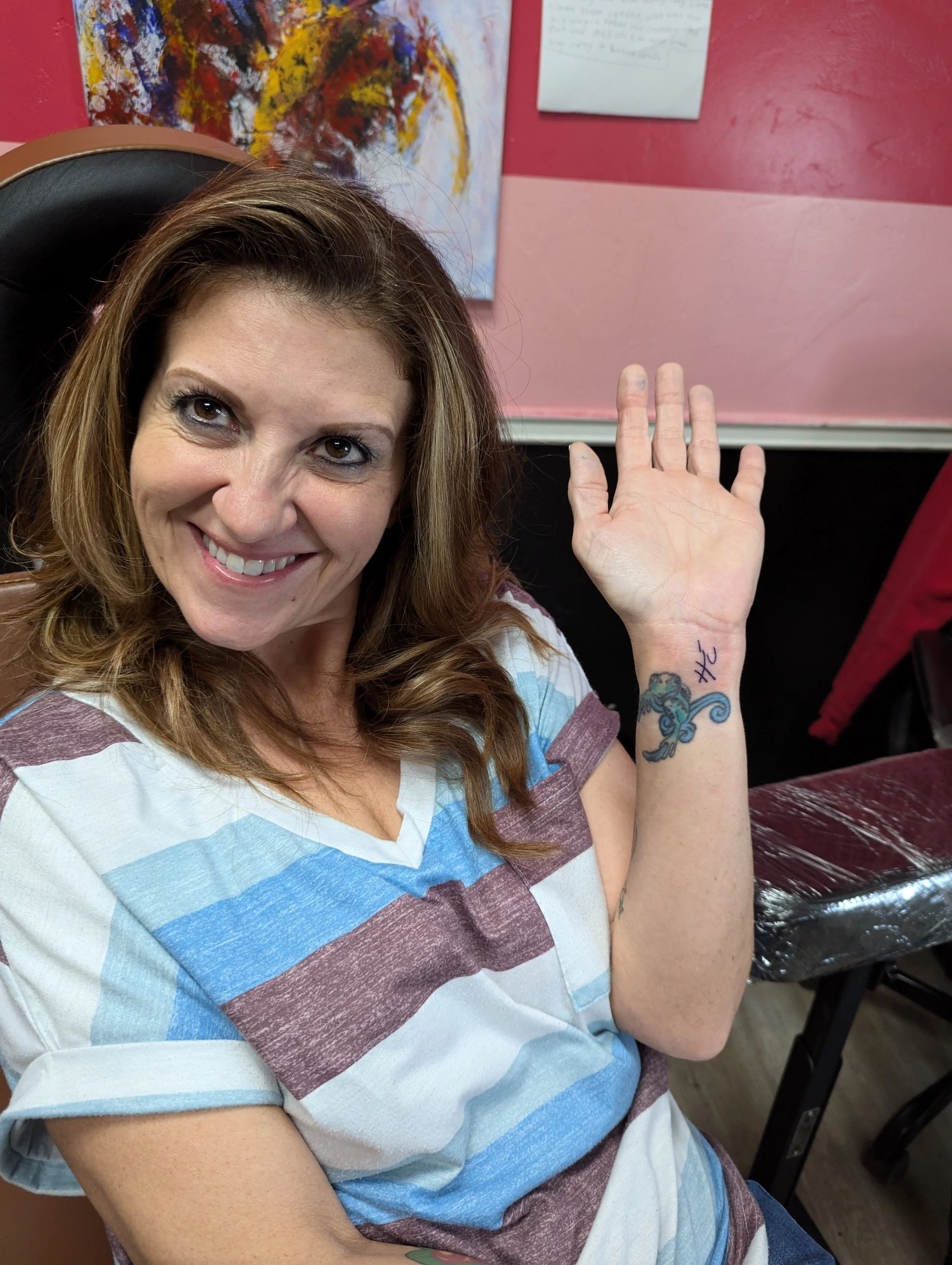 Smiling woman with striped shirt showing a wrist tattoo in a brightly colored room.
