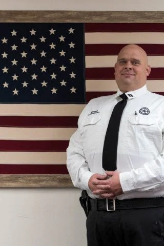 A person in a uniform stands in front of a framed American flag.