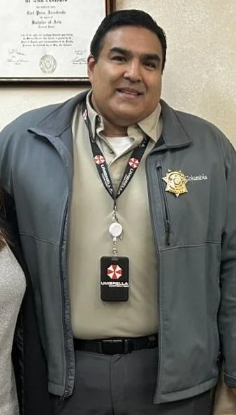 A person wearing a Columbia jacket with a sheriff badge and an Umbrella Corporation lanyard standing indoors, with a document visible on the wall behind.